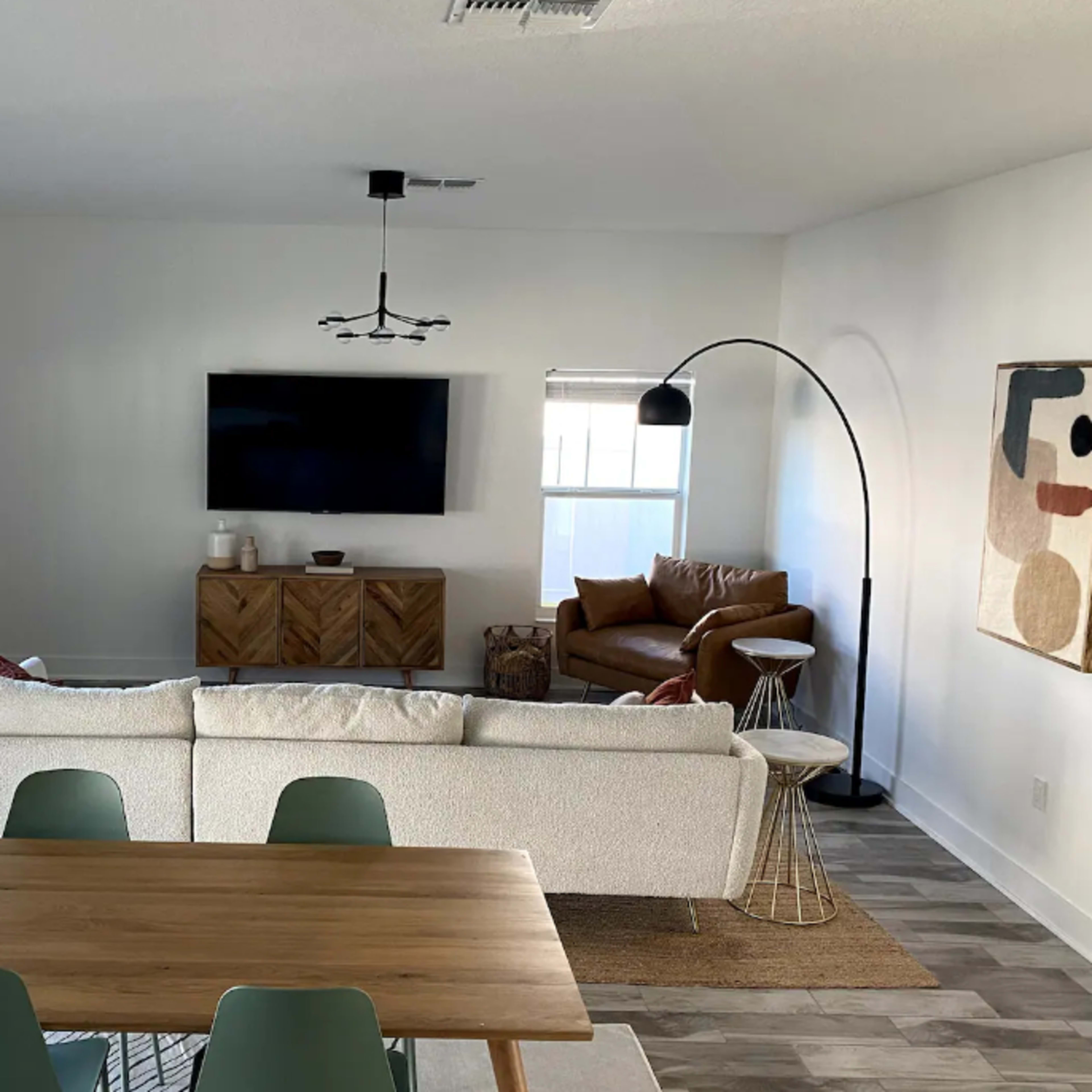 A modern living room features a white sofa, a wooden dining table, a large television on the wall, and a brown leather chair.