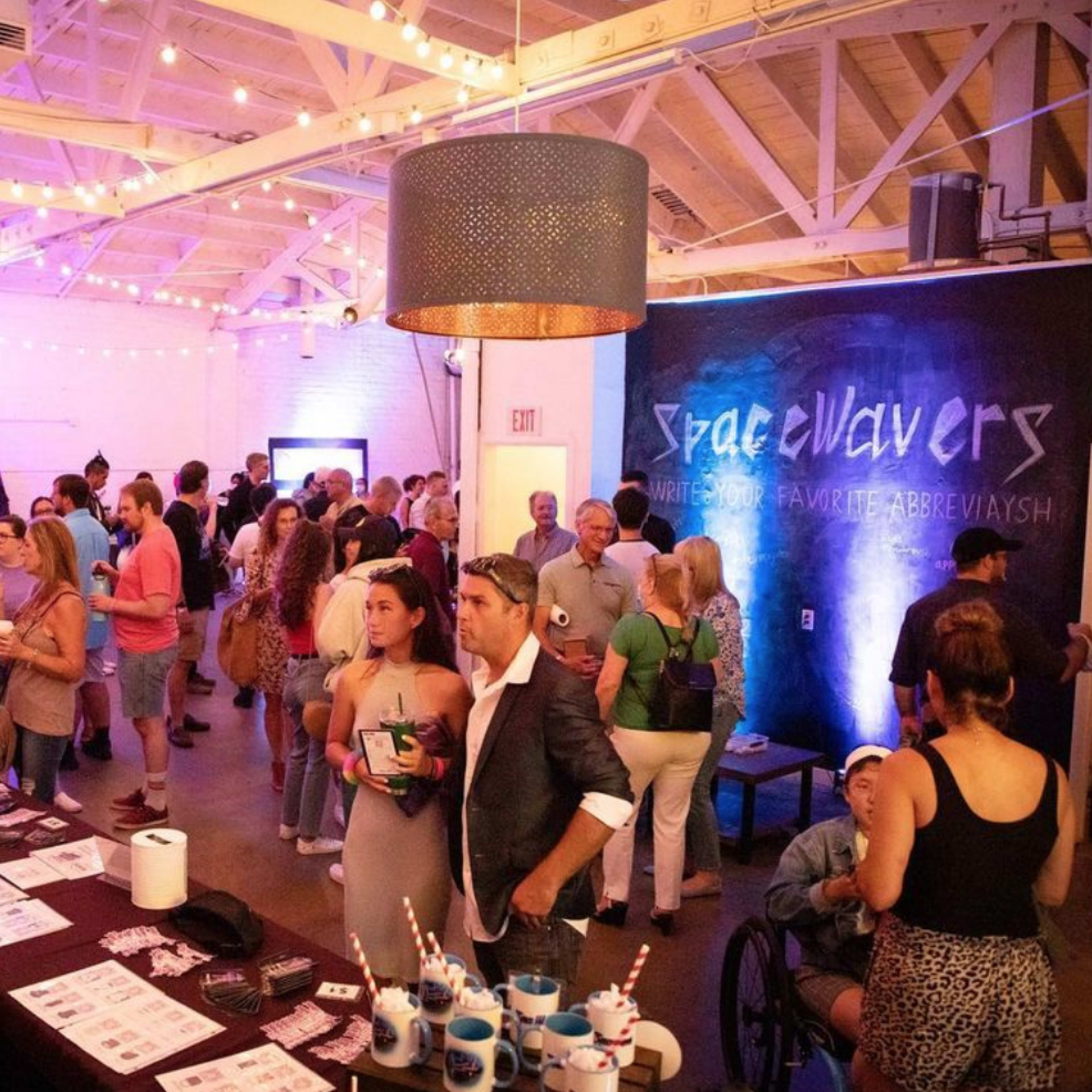 A crowded event space features people mingling, with tables displaying promotional materials and colorful lighting illuminating the room.