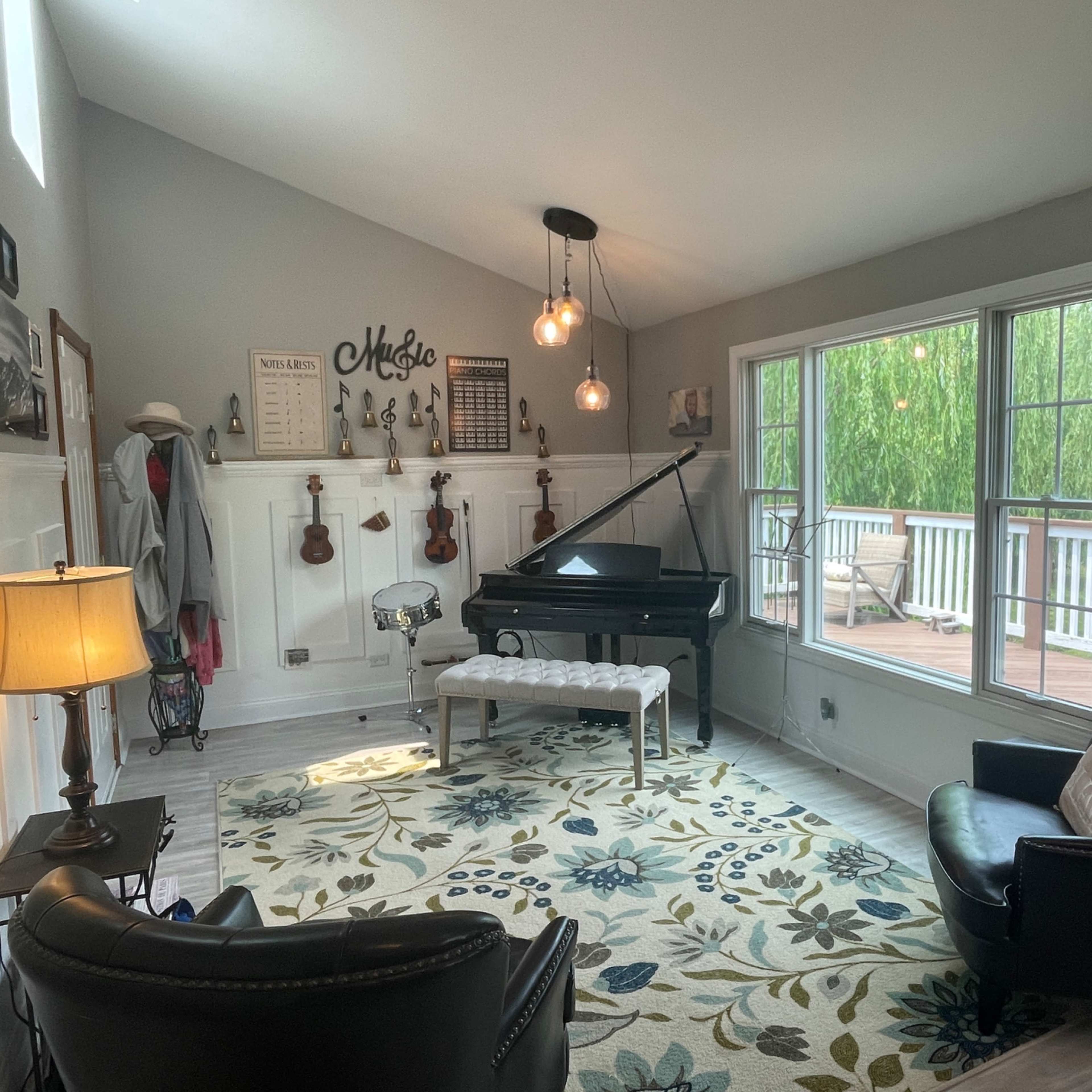 The room features a black piano, a drum set, and various musical instruments displayed on the walls, with two black chairs and a large window overlooking a deck.