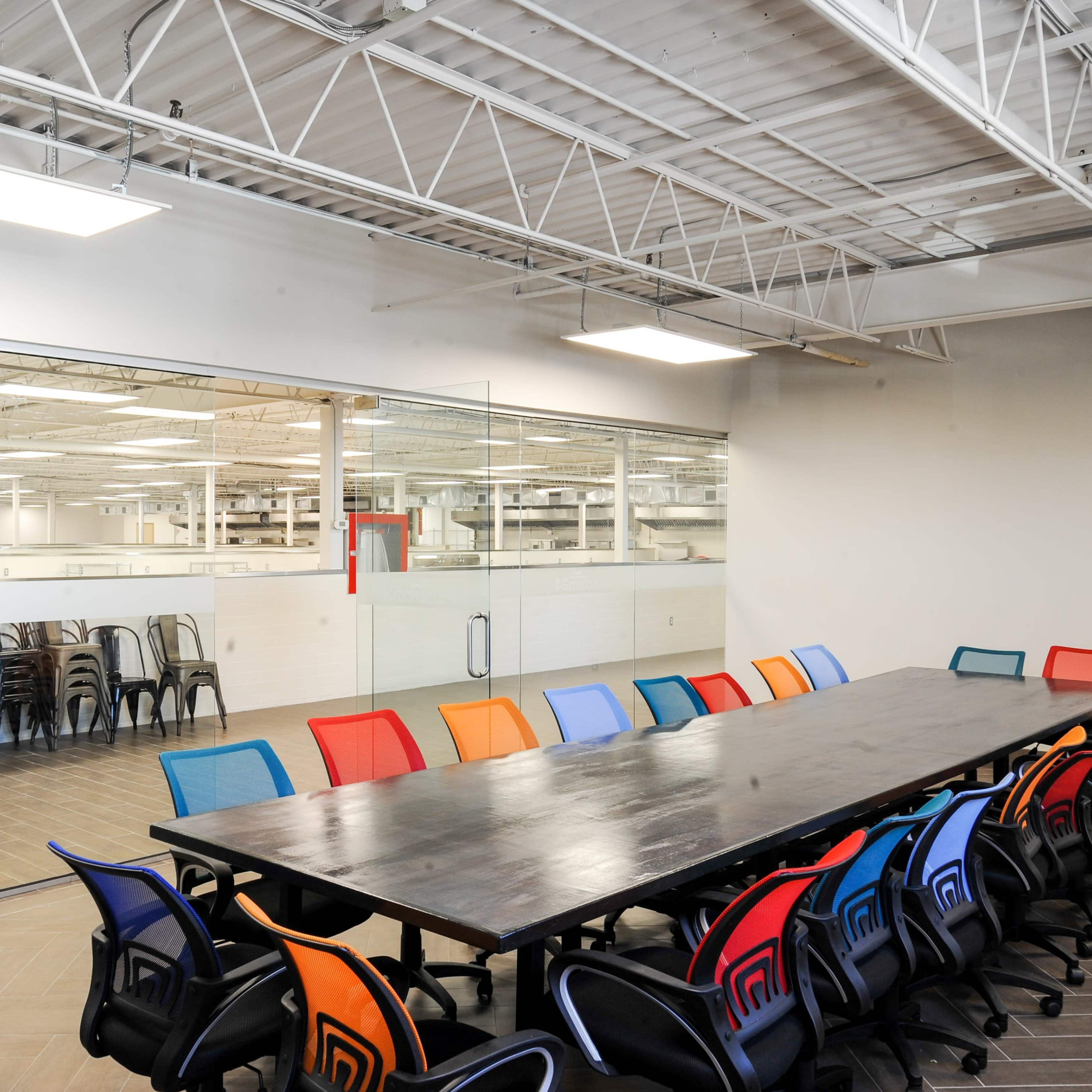 A long, dark wooden conference table is surrounded by colorful chairs in a well-lit office meeting room, with a glass wall revealing another workspace in the background.