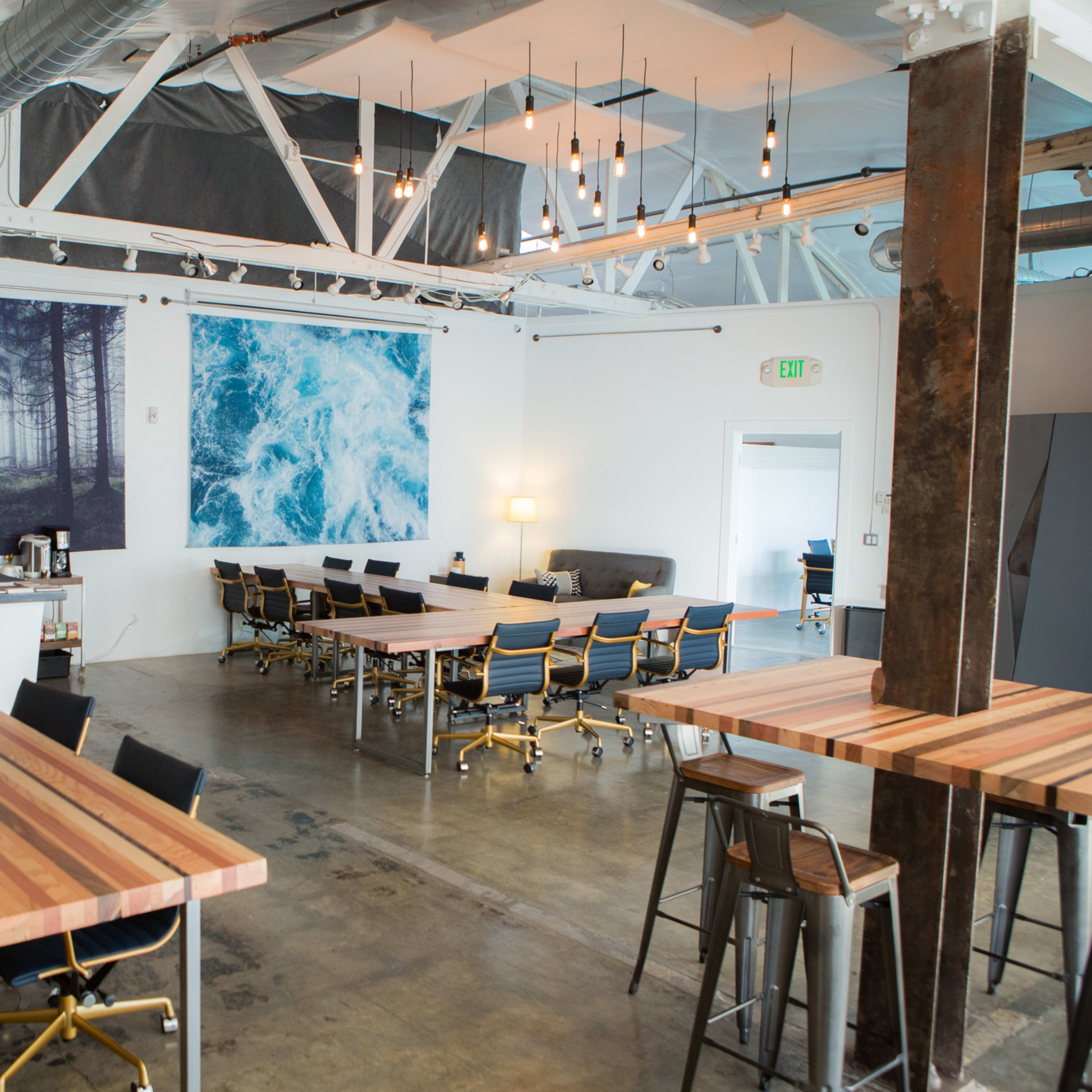A modern workspace with wooden tables, metal chairs, and large wall art depicting a forest and ocean.
