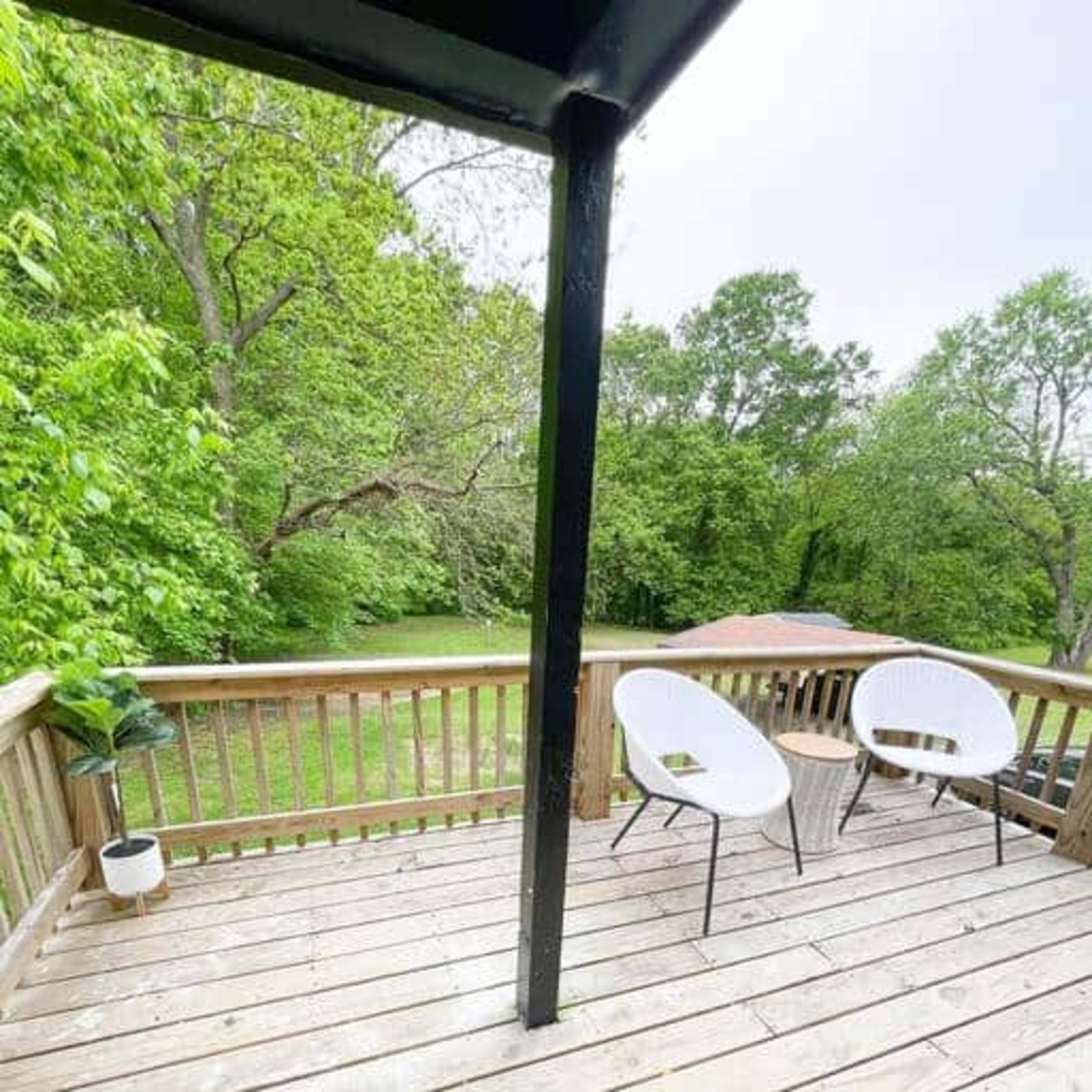 A wooden deck features two white chairs and greenery in the background.