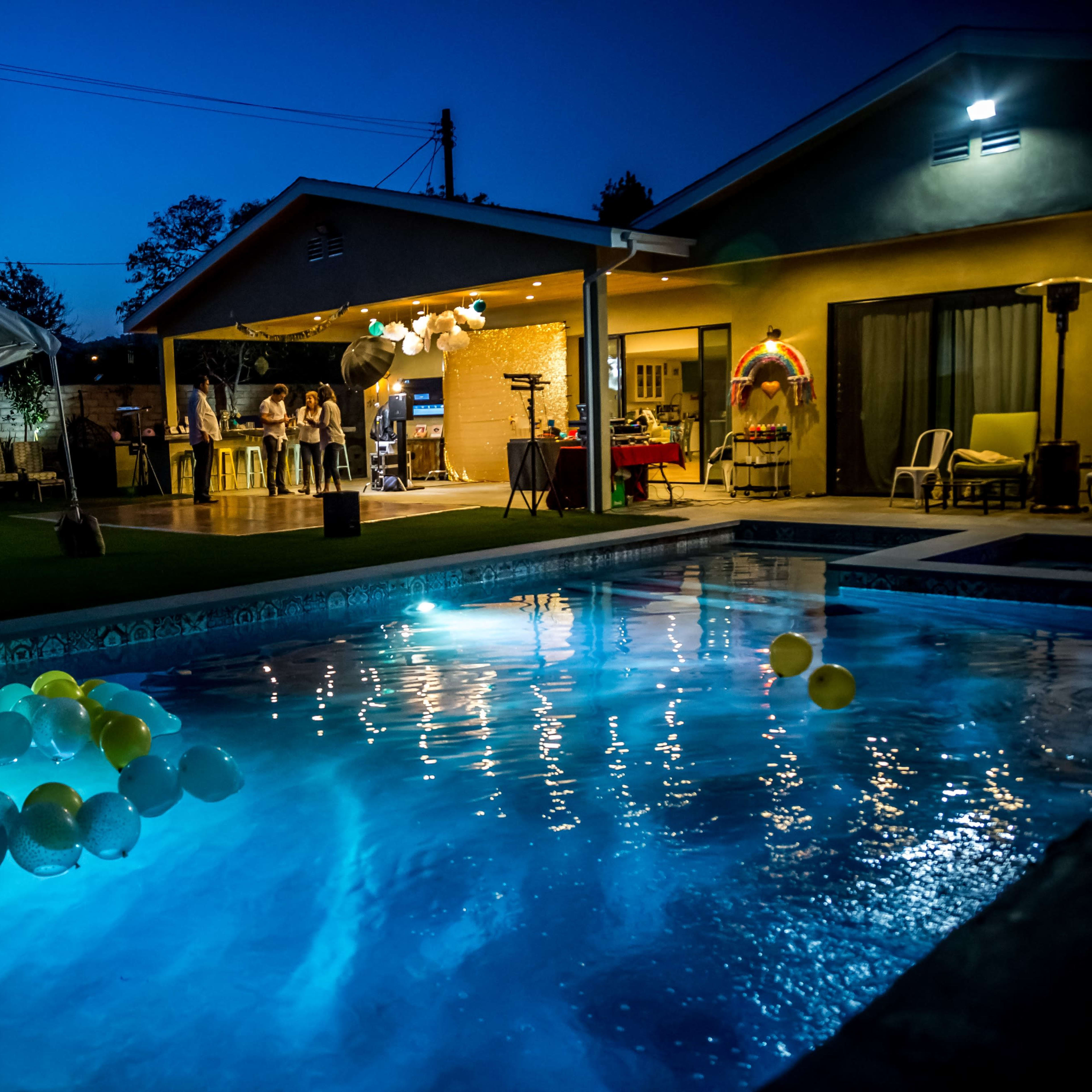 A backyard party scene features a pool illuminated by balloons and a large tent set up next to a house at twilight.