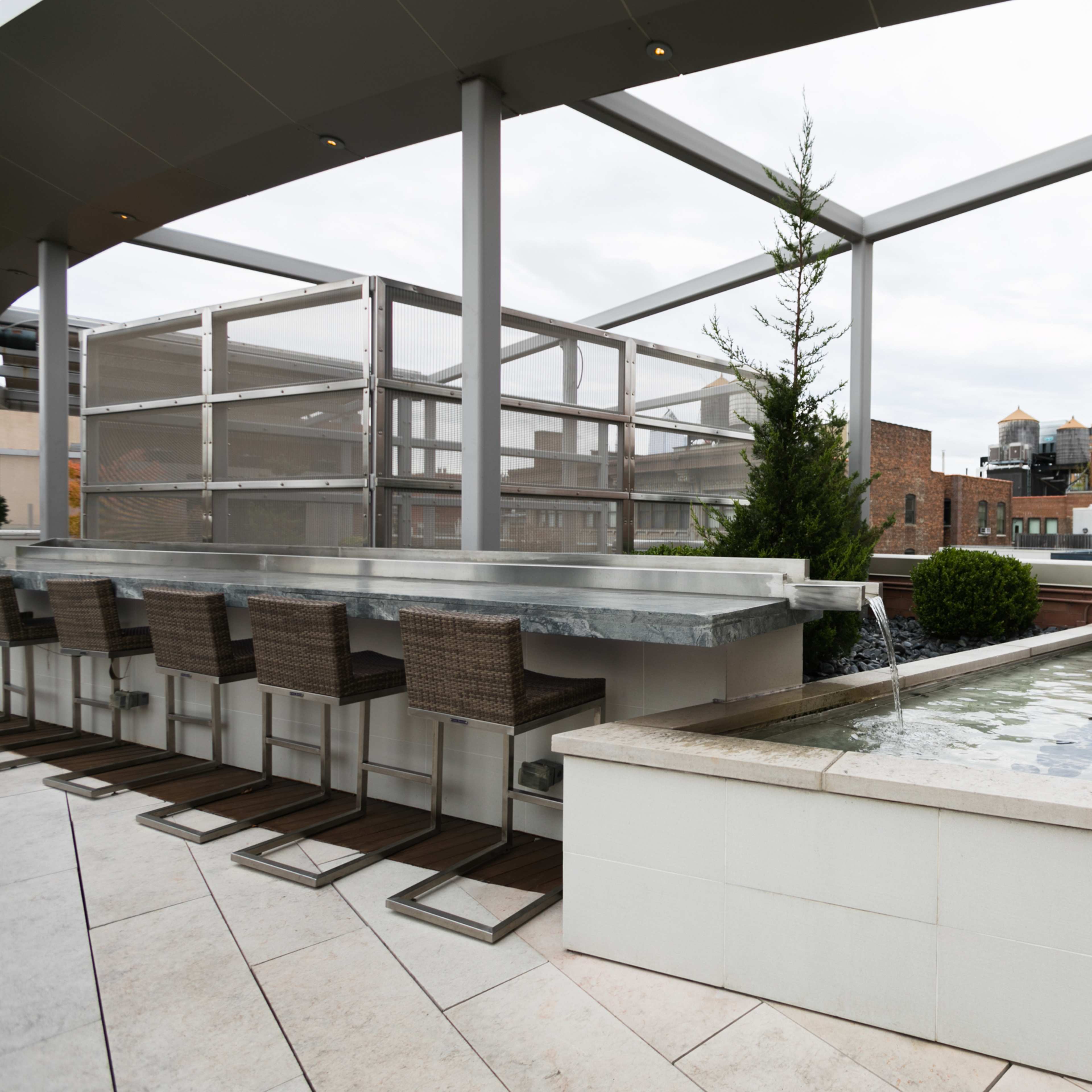 A modern rooftop terrace featuring a bar area with seating and a rectangular water fountain amid urban buildings in the background.