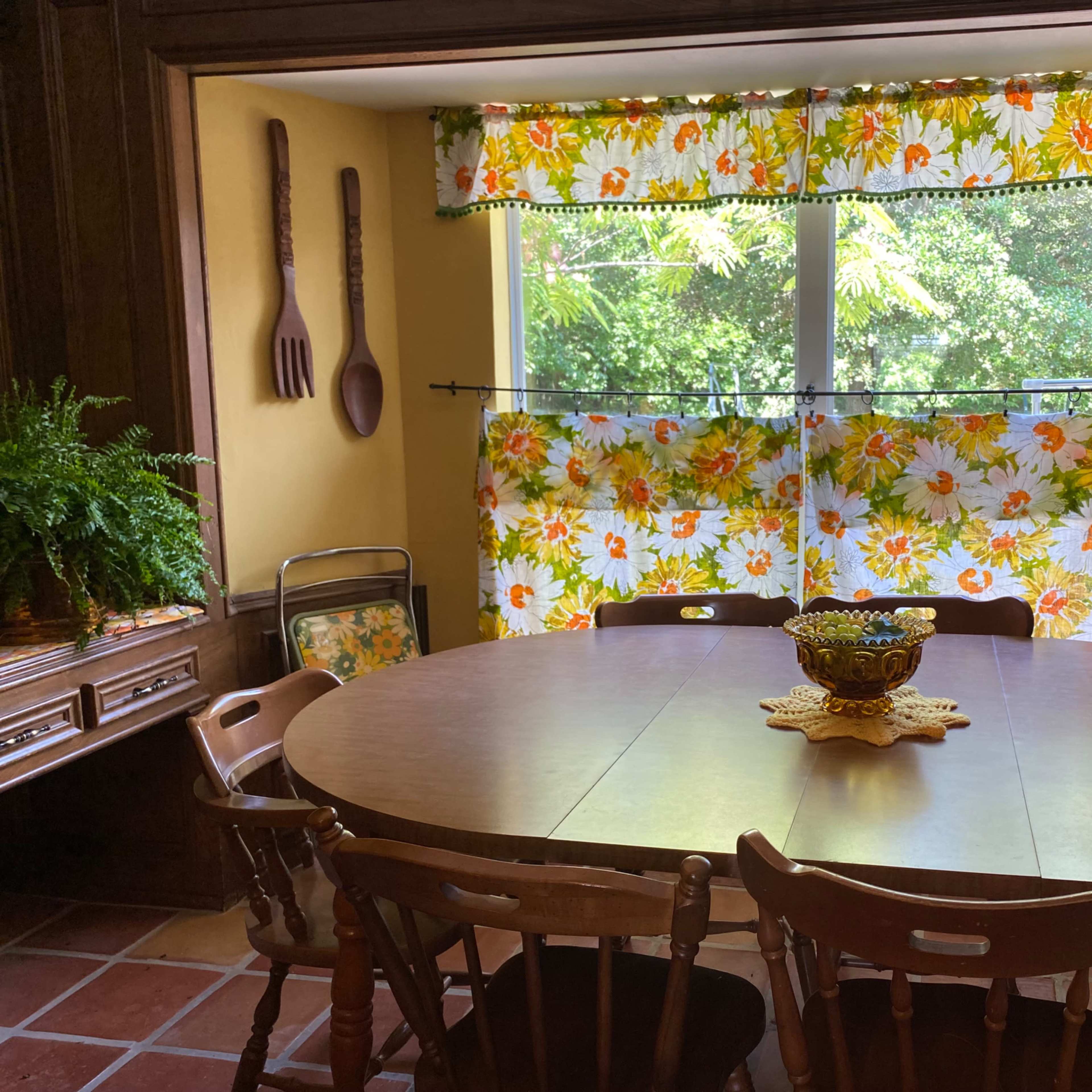A dining room features a round wooden table surrounded by chairs, with floral-patterned curtains covering a window that overlooks greenery.
