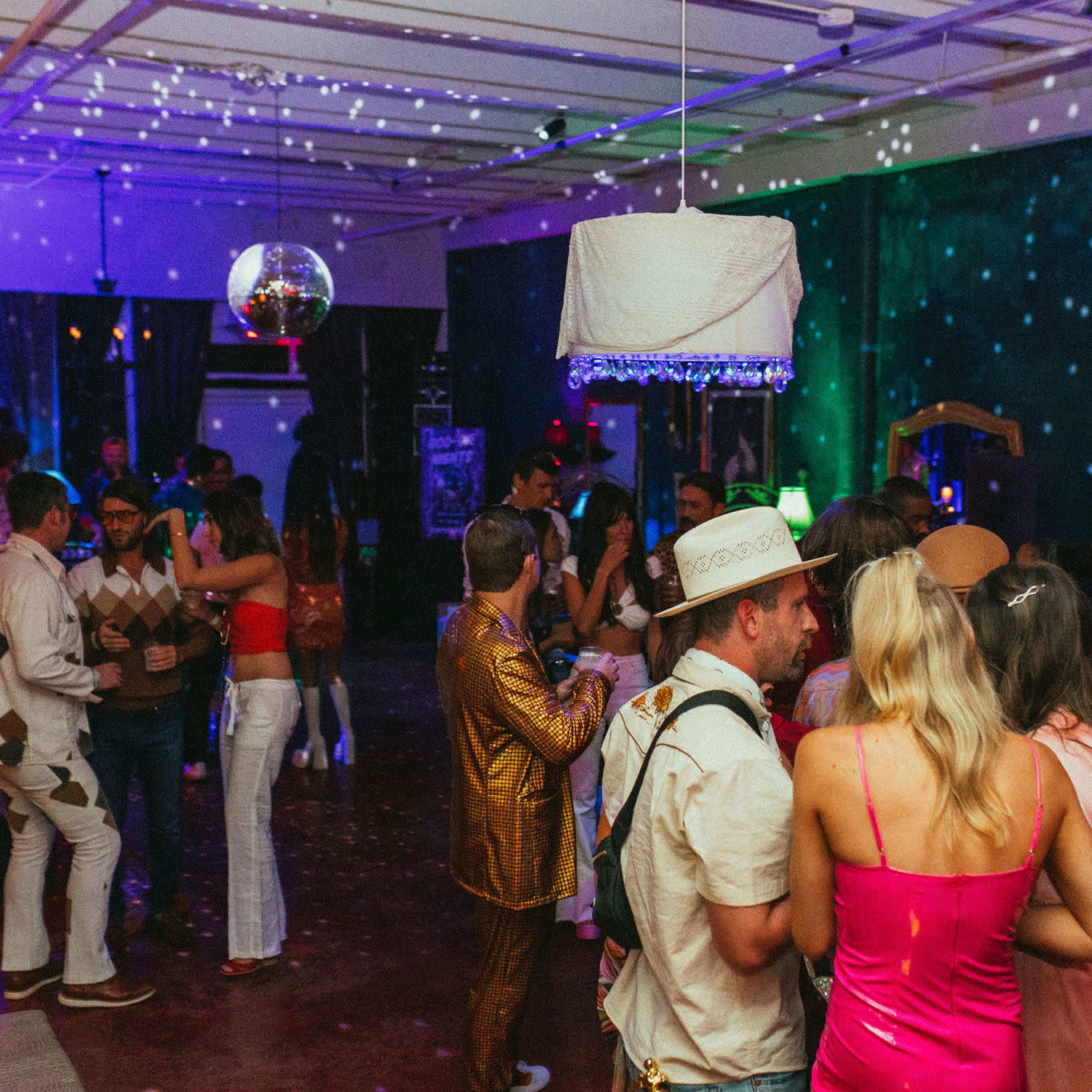 A crowded indoor party features attendees in various costumes and colorful decorations, with a disco ball reflecting light across the room.