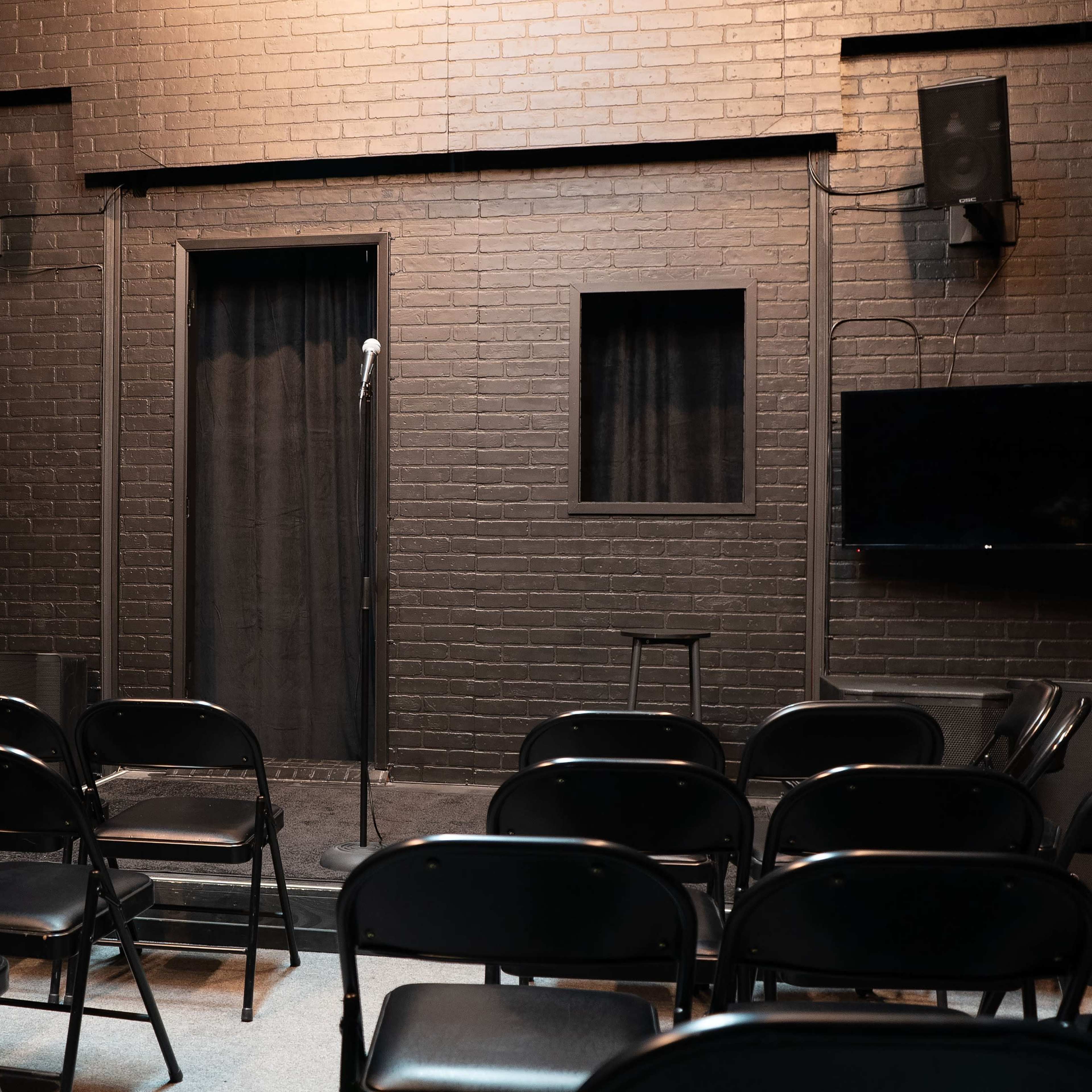 A dimly lit comedy stage features a microphone stand and a backdrop of black brick, with rows of black folding chairs arranged in front.