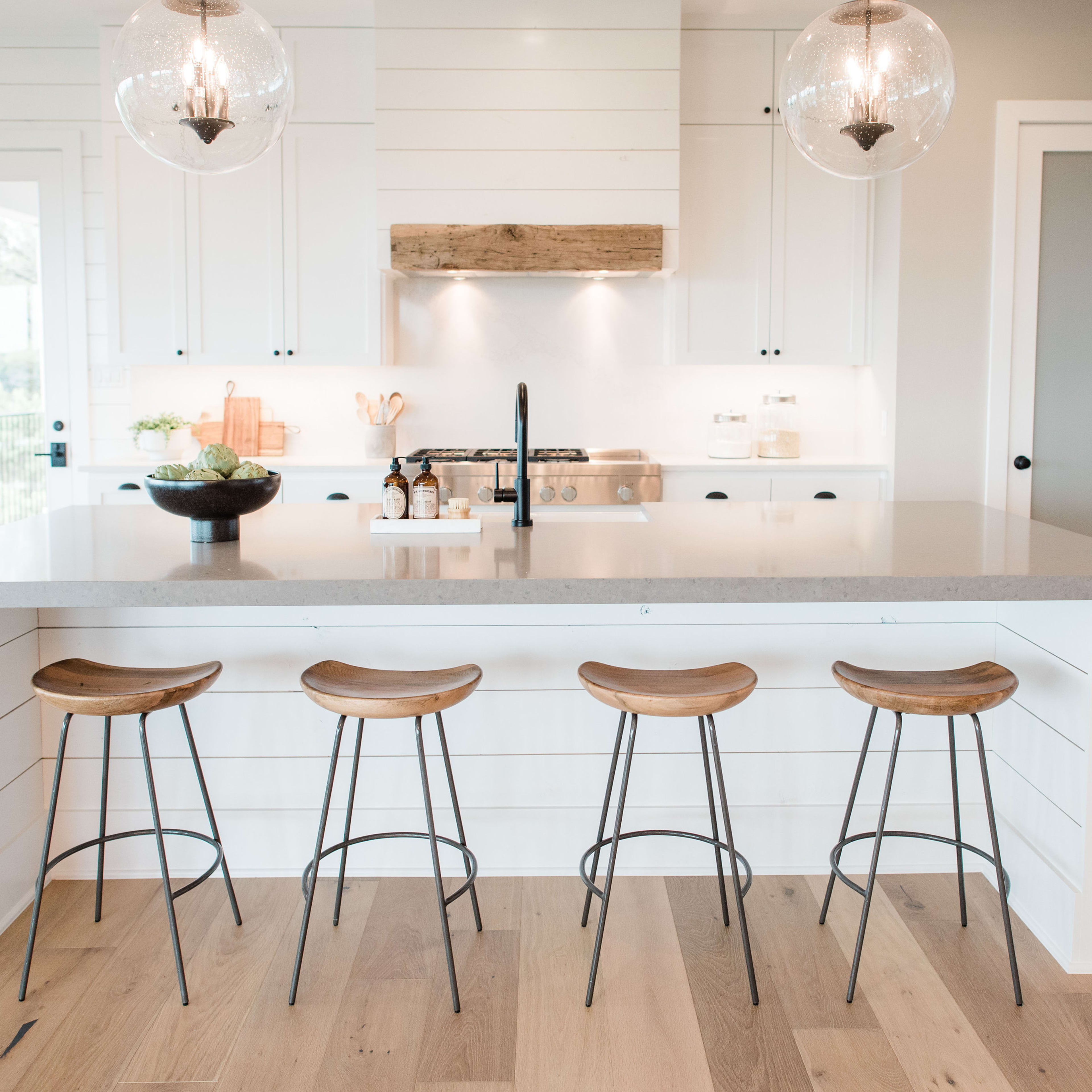 A modern kitchen featuring a long countertop, four wooden stools, and pendant lighting over a cooking area.