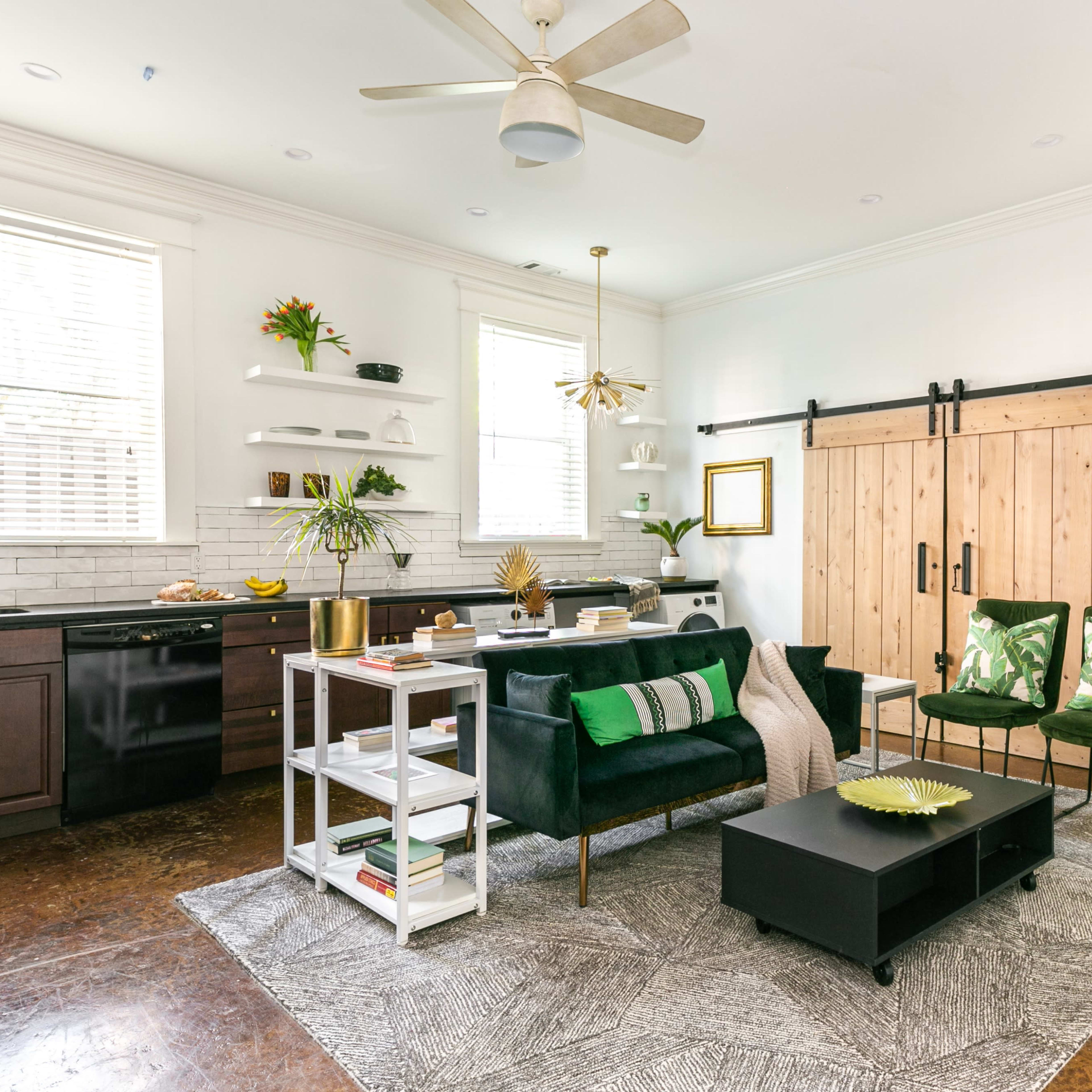 A modern living room and kitchen area featuring a dark green sofa, a black coffee table, a sliding barn door, and a kitchen with dark cabinets and open shelving.