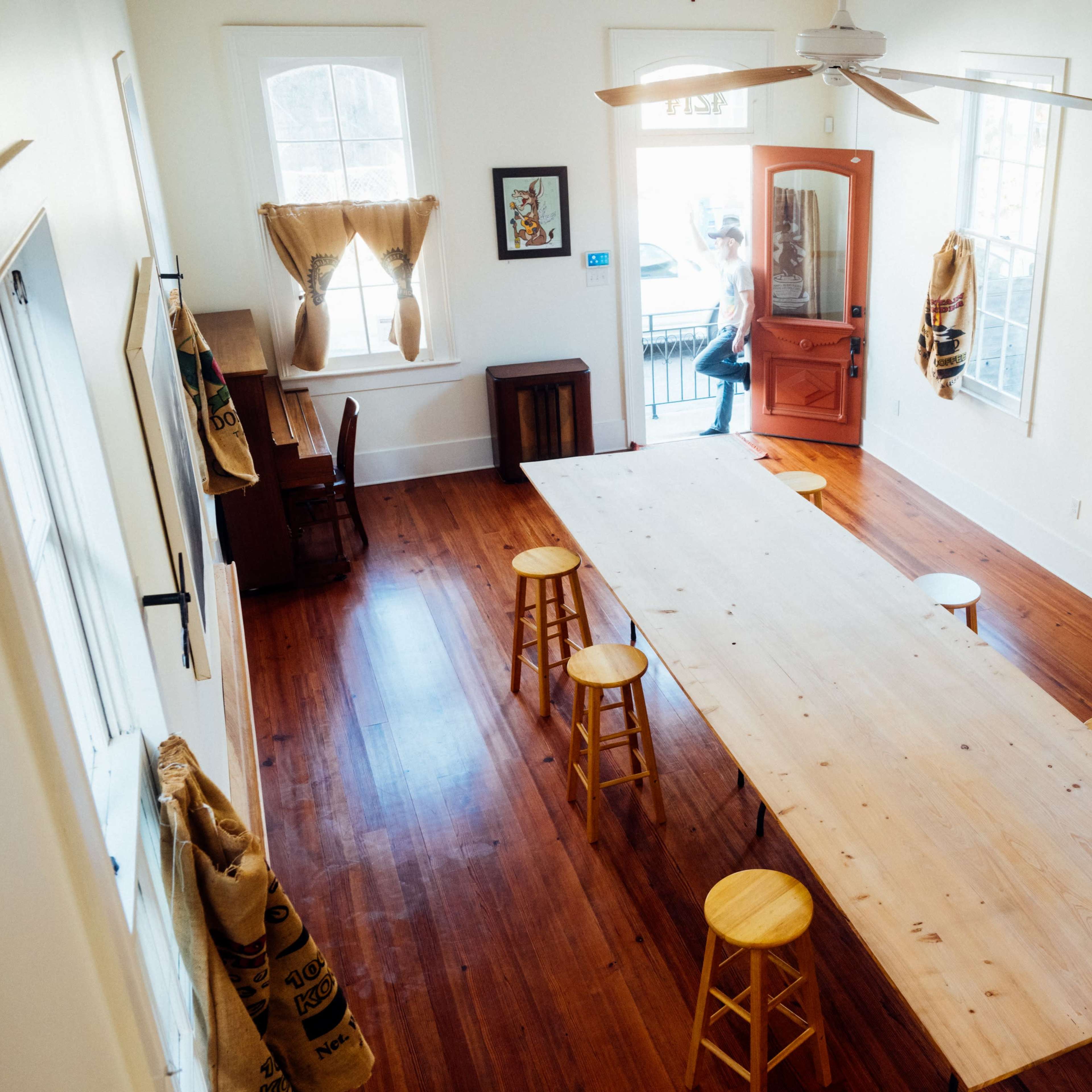 A spacious, well-lit room features a long wooden table surrounded by stools, with a person exiting through an open door.