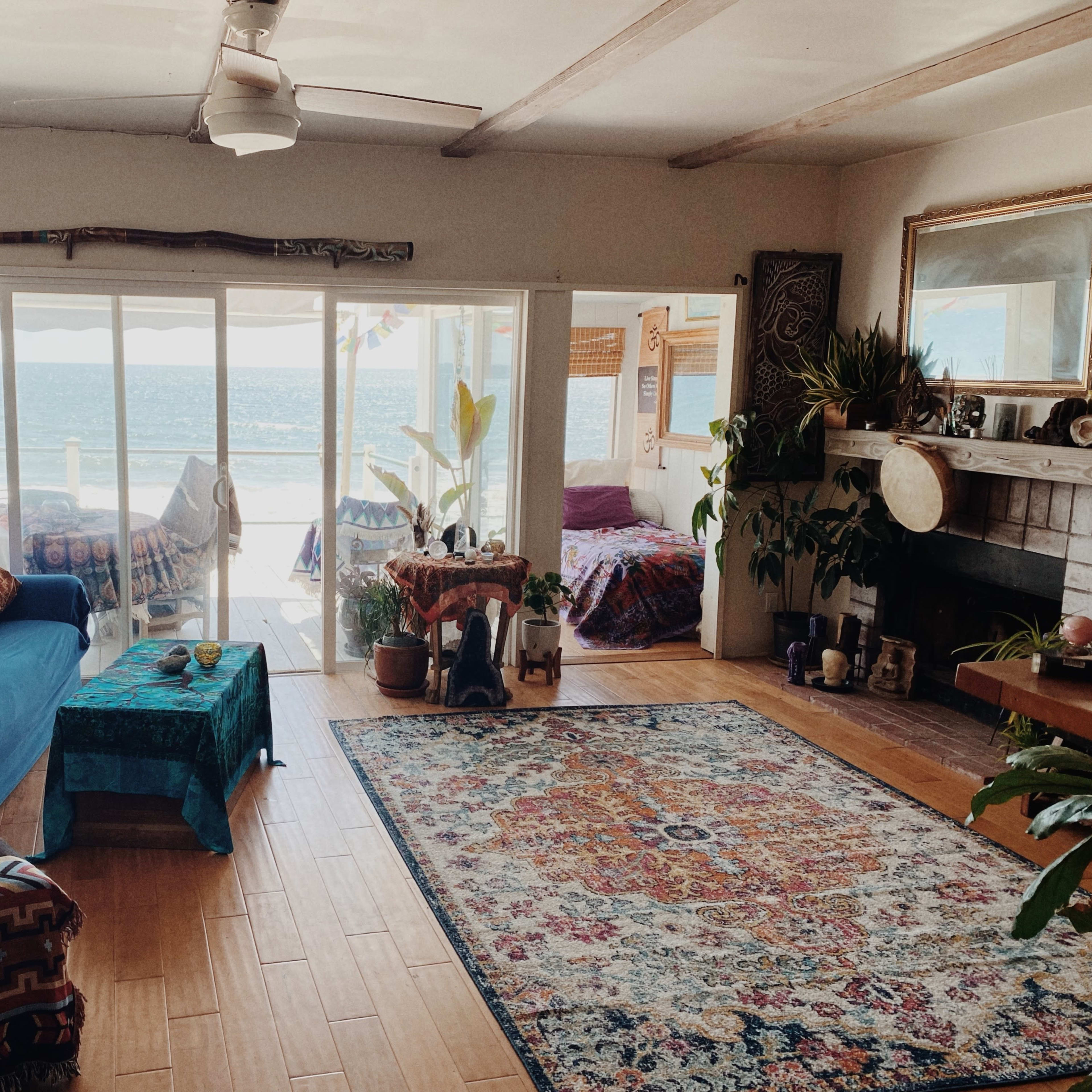 A cozy living room features a blue couch, colorful patterned rugs, plants, and large windows overlooking the ocean.