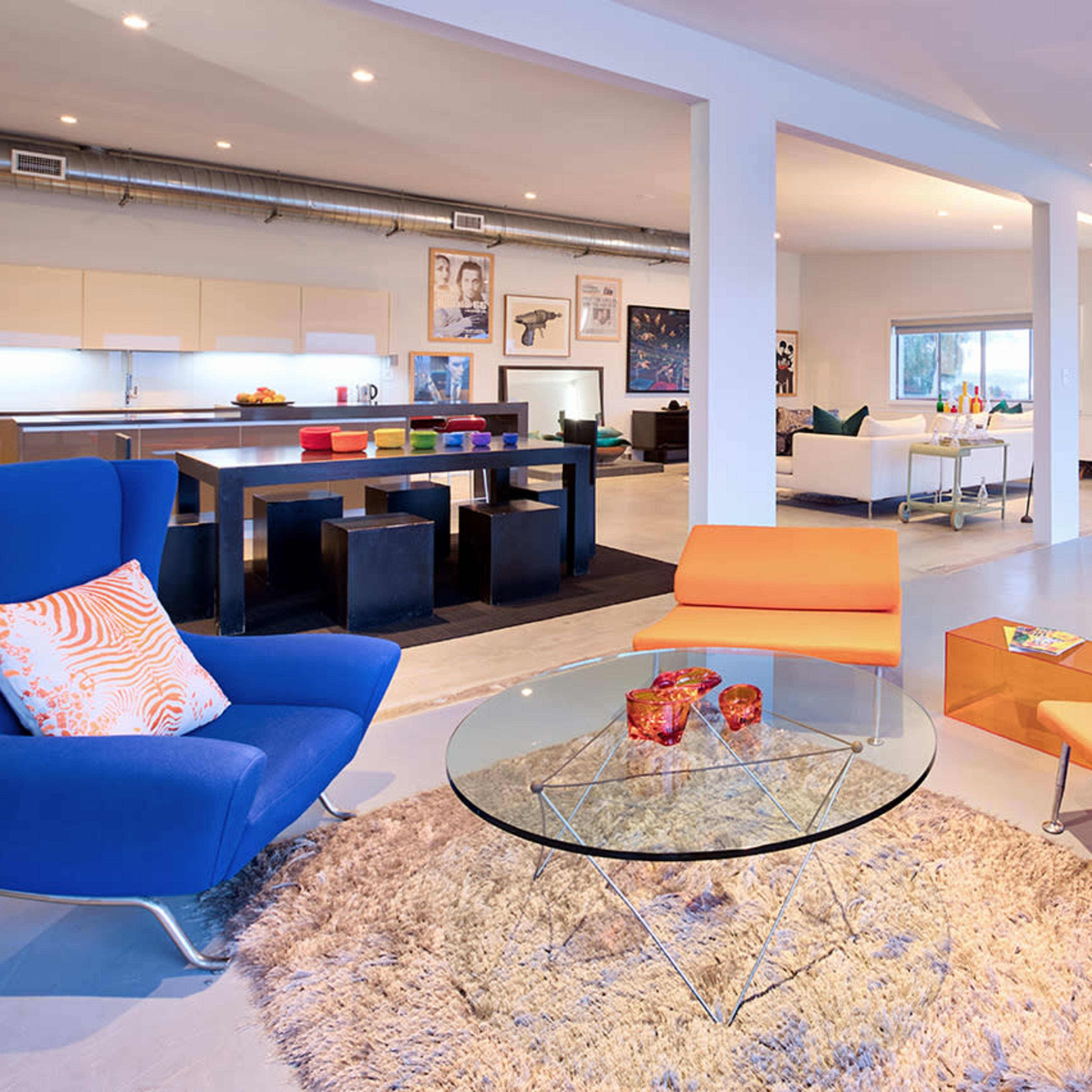 A modern living space with colorful furniture, including a blue chair and an orange couch, arranged around a glass coffee table on a large rug.