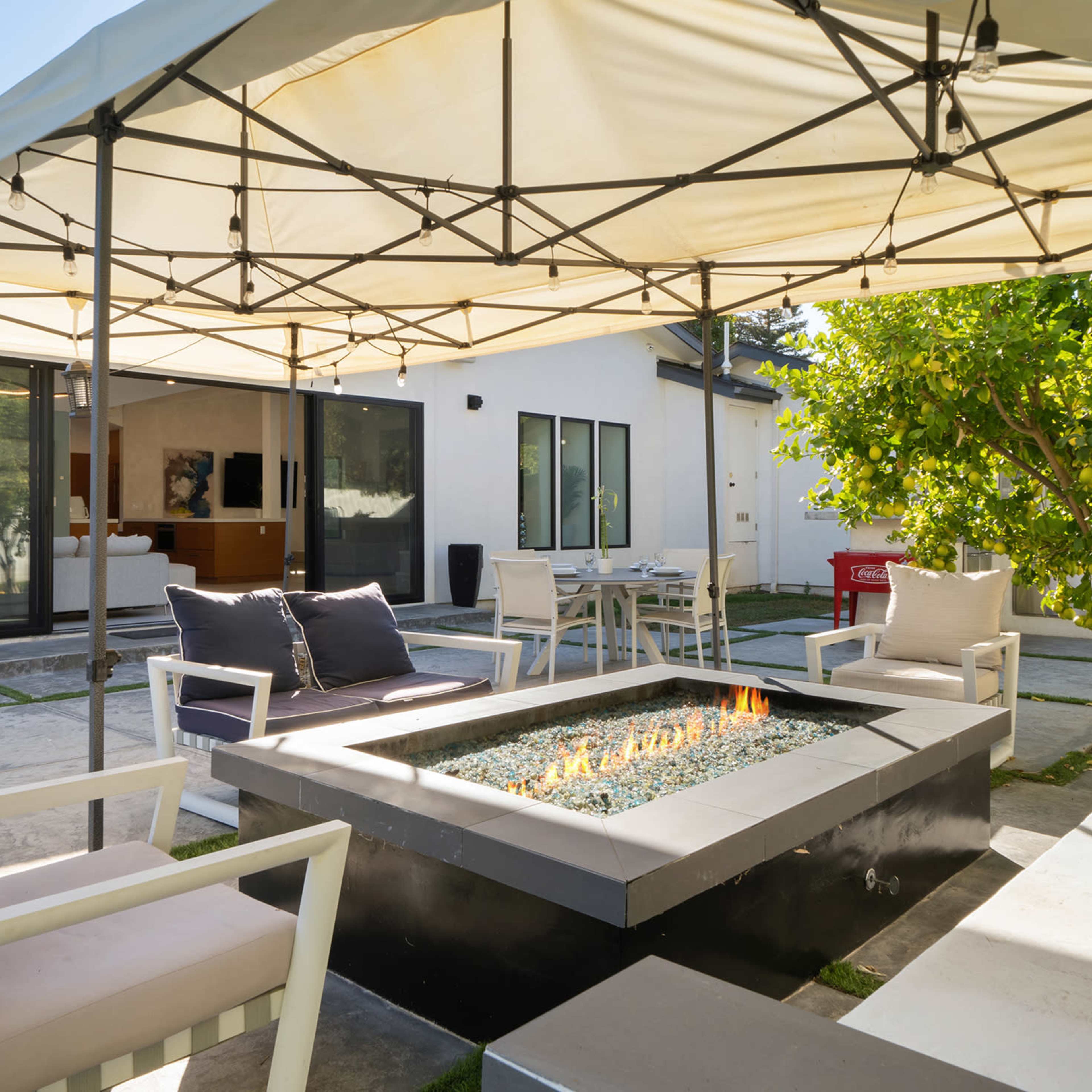 A patio area features a fire pit surrounded by seating and a canopy, with a view of a modern home in the background.