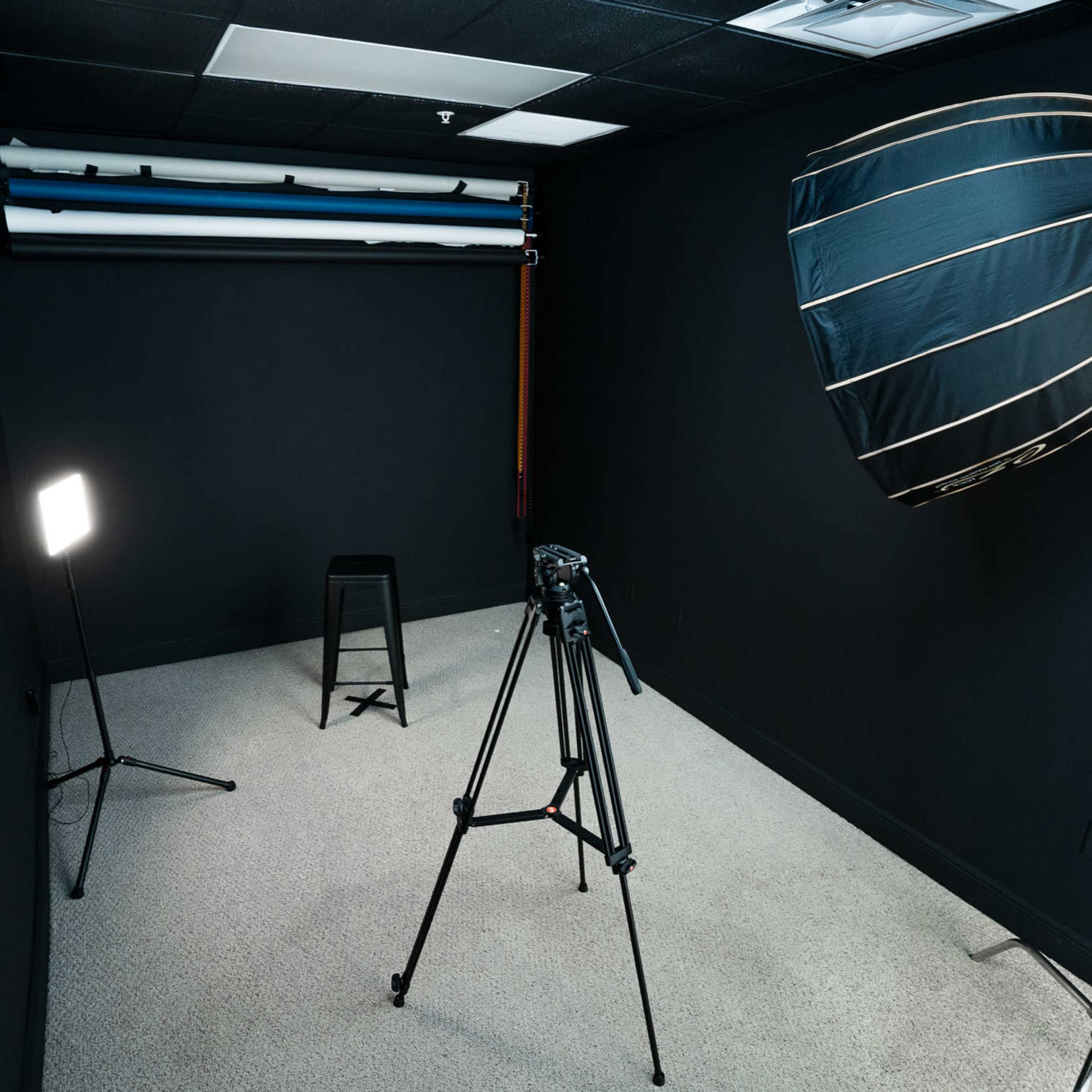 A photography studio with a black backdrop, a stool, a camera tripod, and lighting equipment.