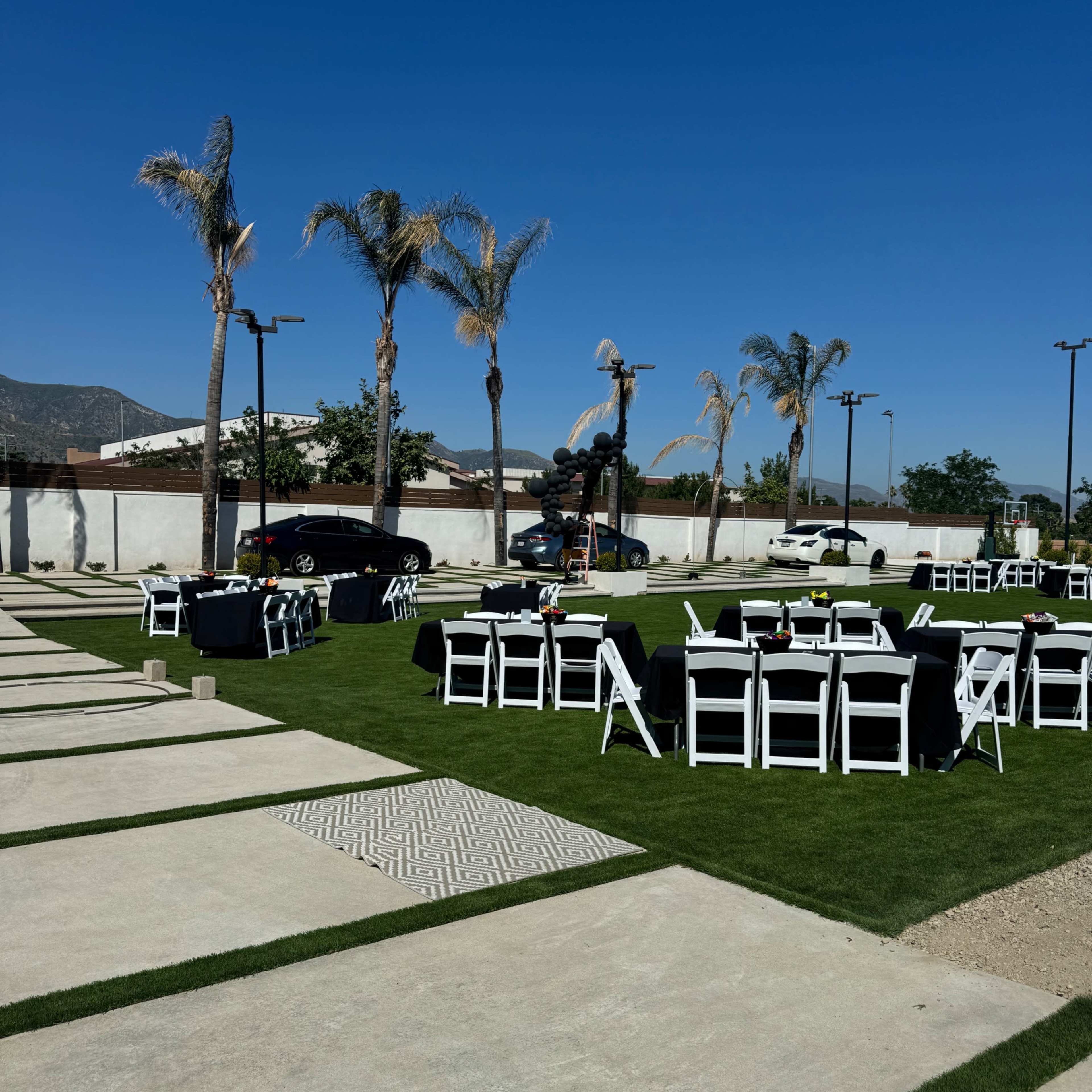 The scene features a landscaped outdoor area set with black and white tables and chairs, surrounded by palm trees and a clear blue sky.