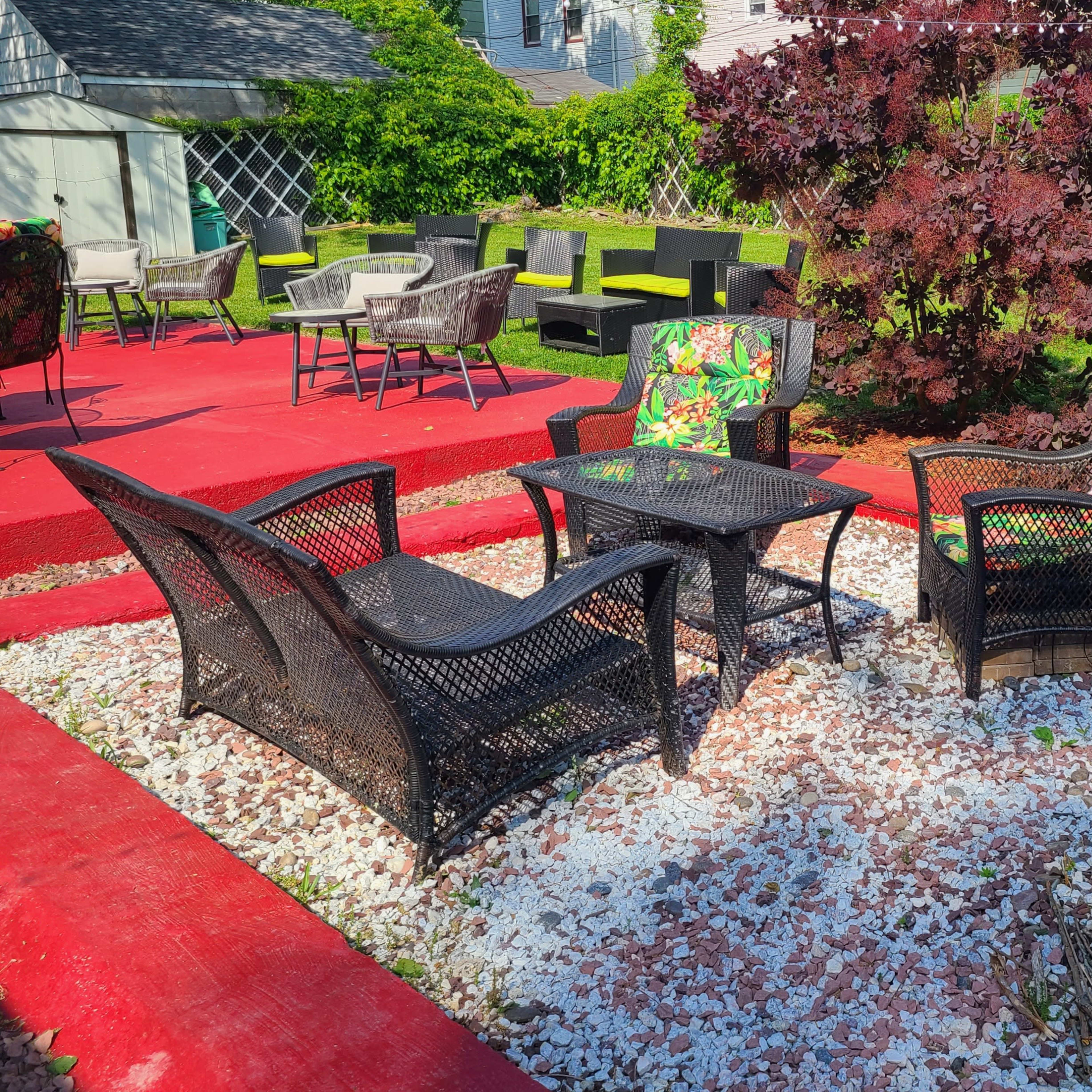 A patio area with four black wicker chairs and a small table arranged around a fire pit, surrounded by gravel and vibrant red landscaping.