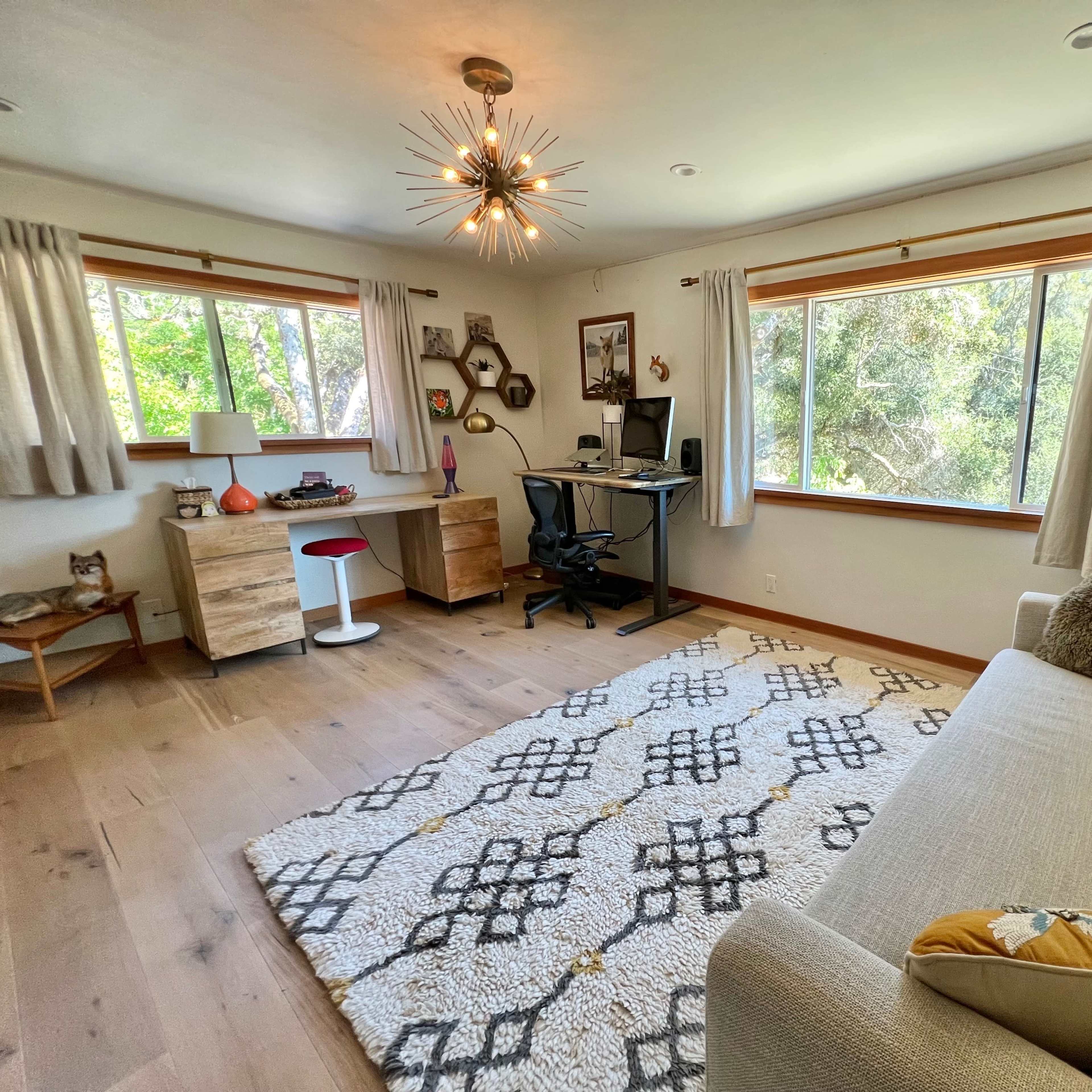 A bright room features a sofa, a desk with a computer, and large windows overlooking greenery, with a patterned rug on the wooden floor.