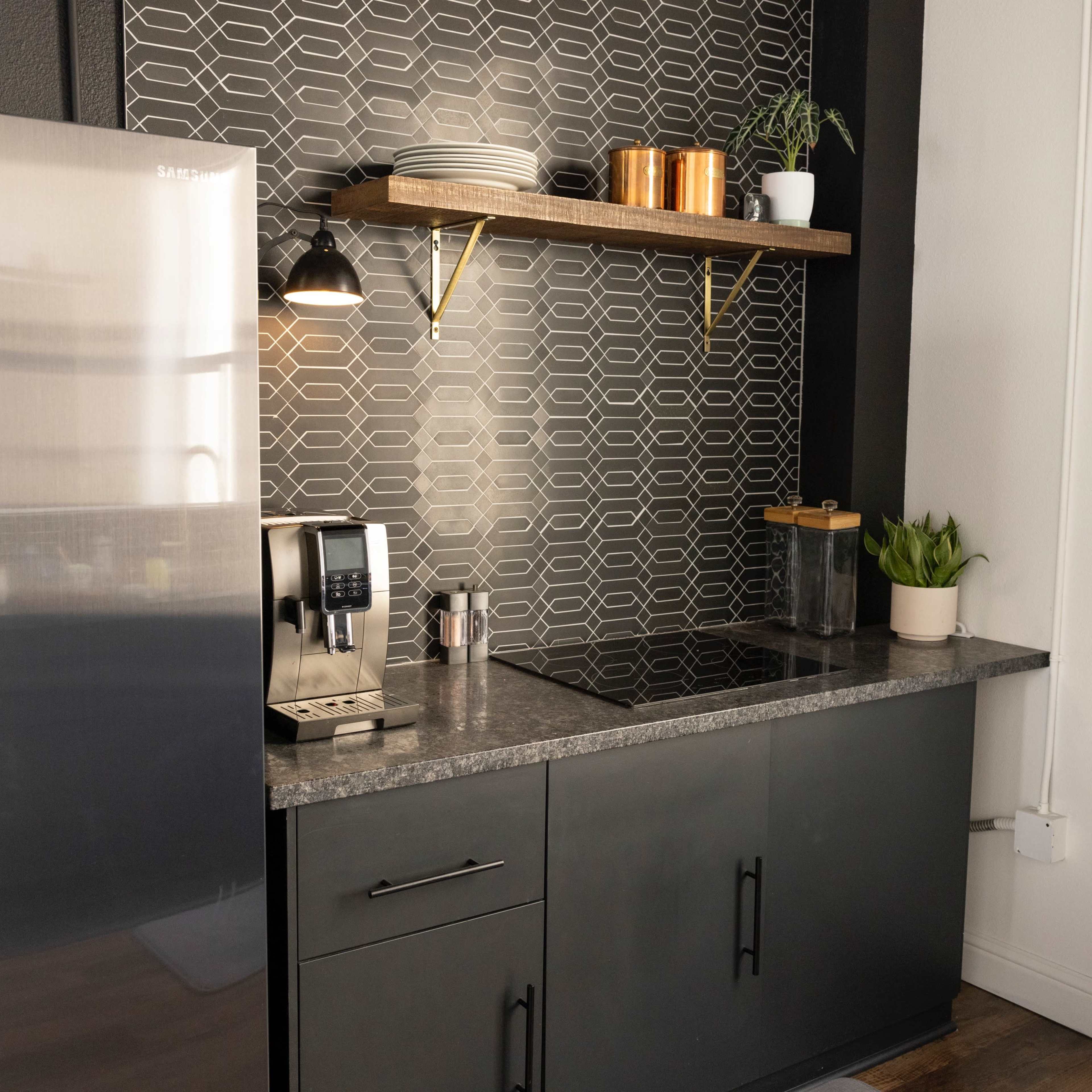 A modern kitchen corner features a stainless steel refrigerator, a sleek countertop with a coffee maker, and open shelving displaying plates and plant decor against a patterned backsplash.
