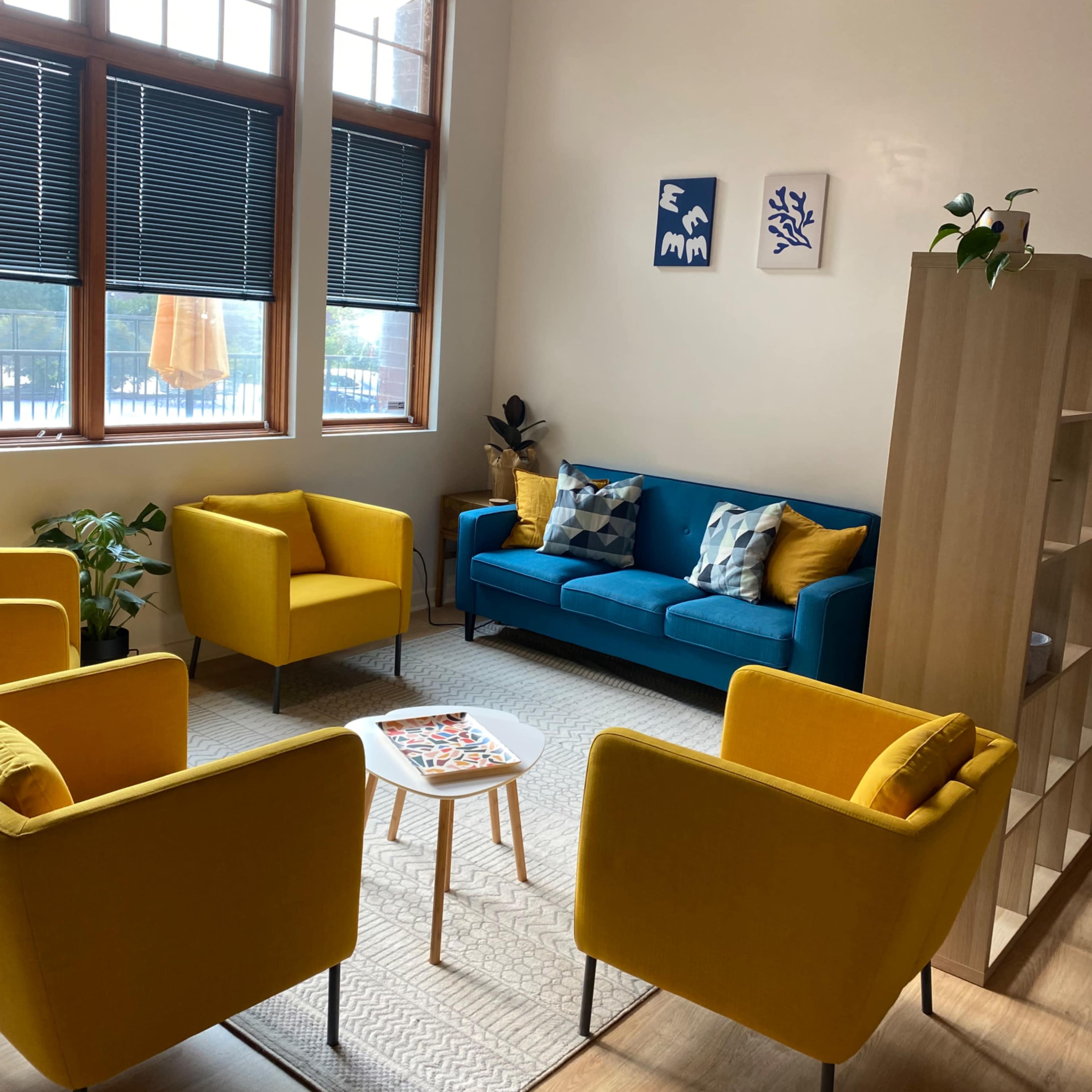 A cozy room with a blue sofa and four yellow chairs arranged around a small table, accented by decorative art on the walls and a plant in the corner.