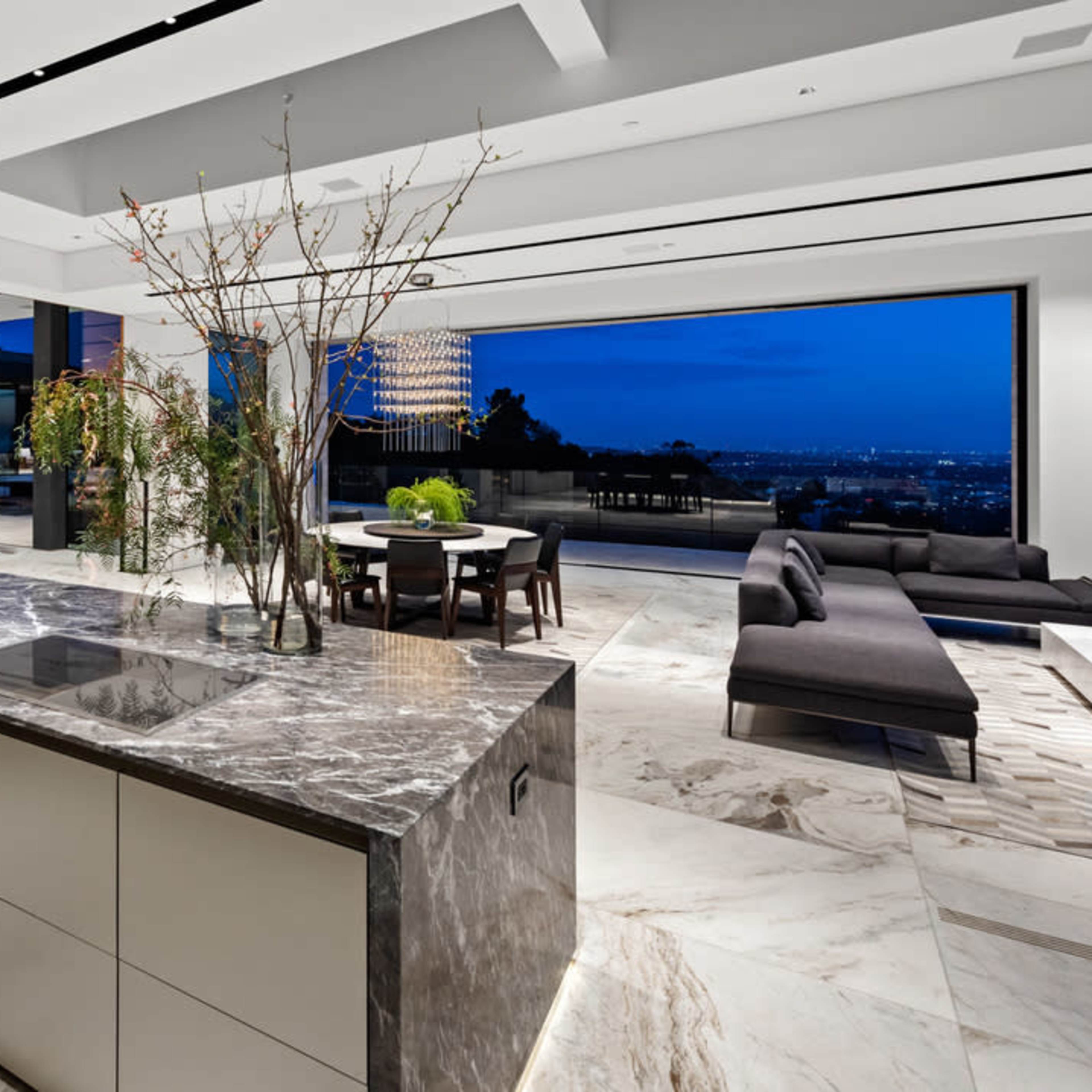 A modern kitchen with a marble island, contemporary decor, and large windows overlooking a city landscape at dusk.