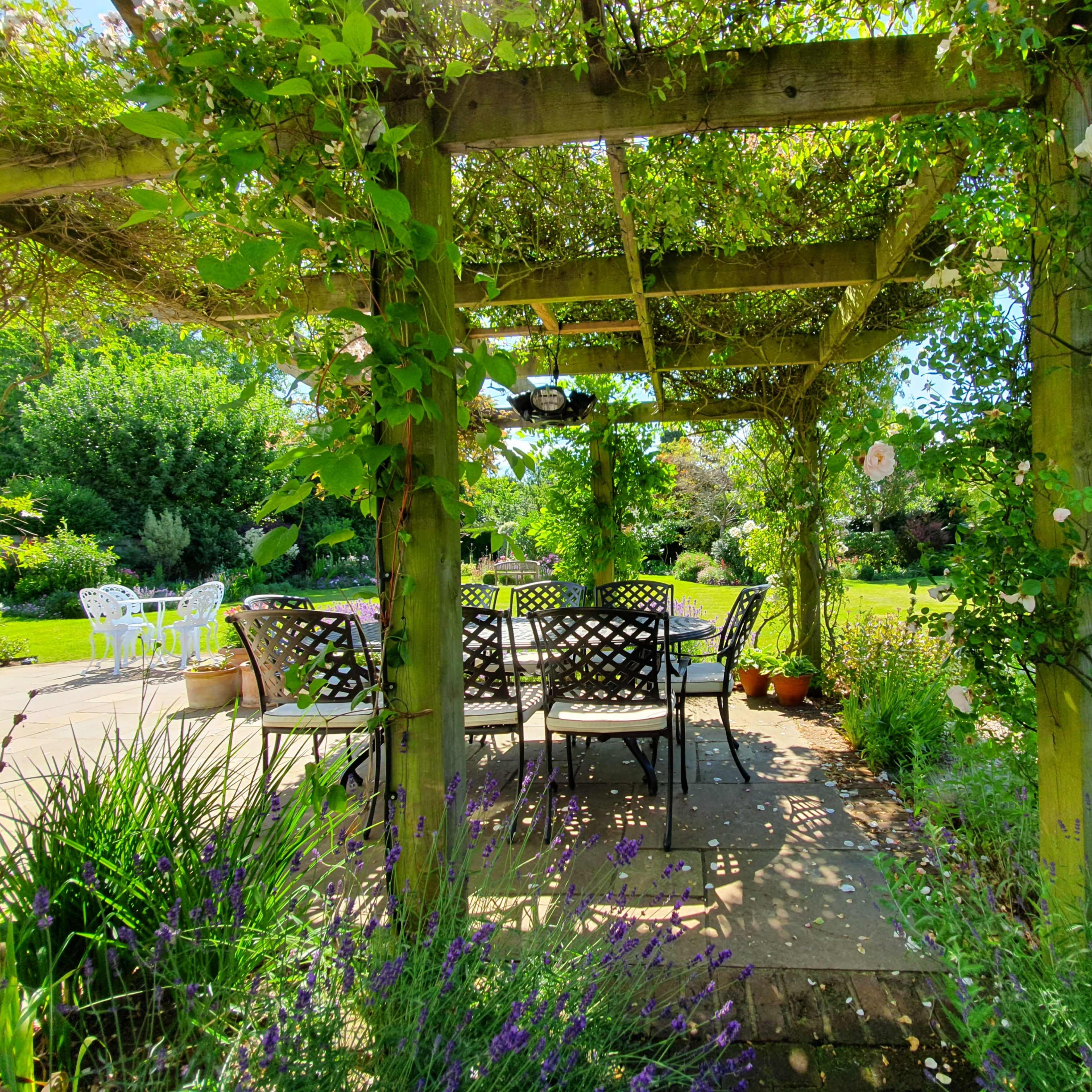 A wooden pergola covered with greenery frames a stone patio with a table and chairs, overlooking a lush garden.