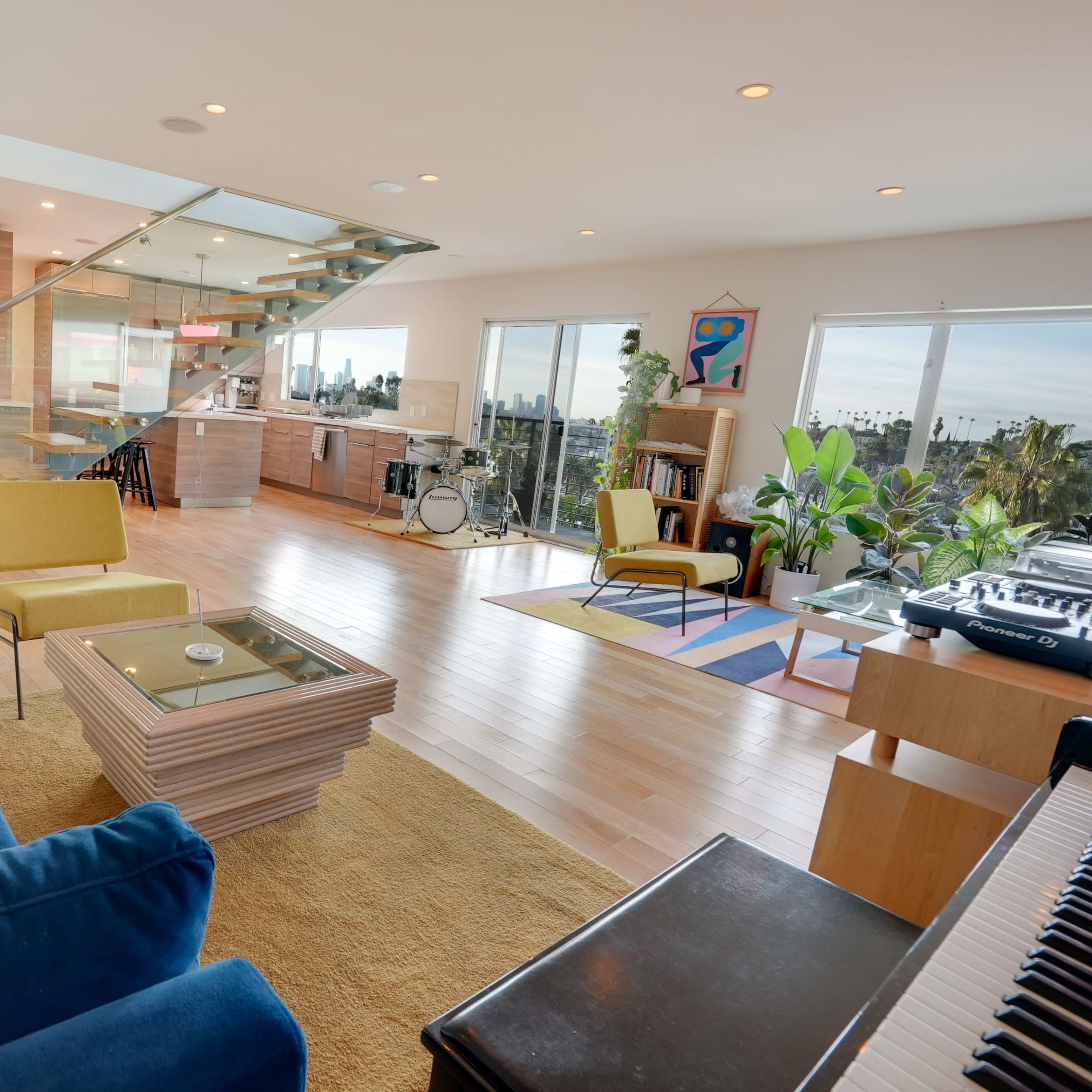 The brightly lit living room features a blue sofa, yellow chairs, a modern coffee table, and large windows showcasing a panoramic view.