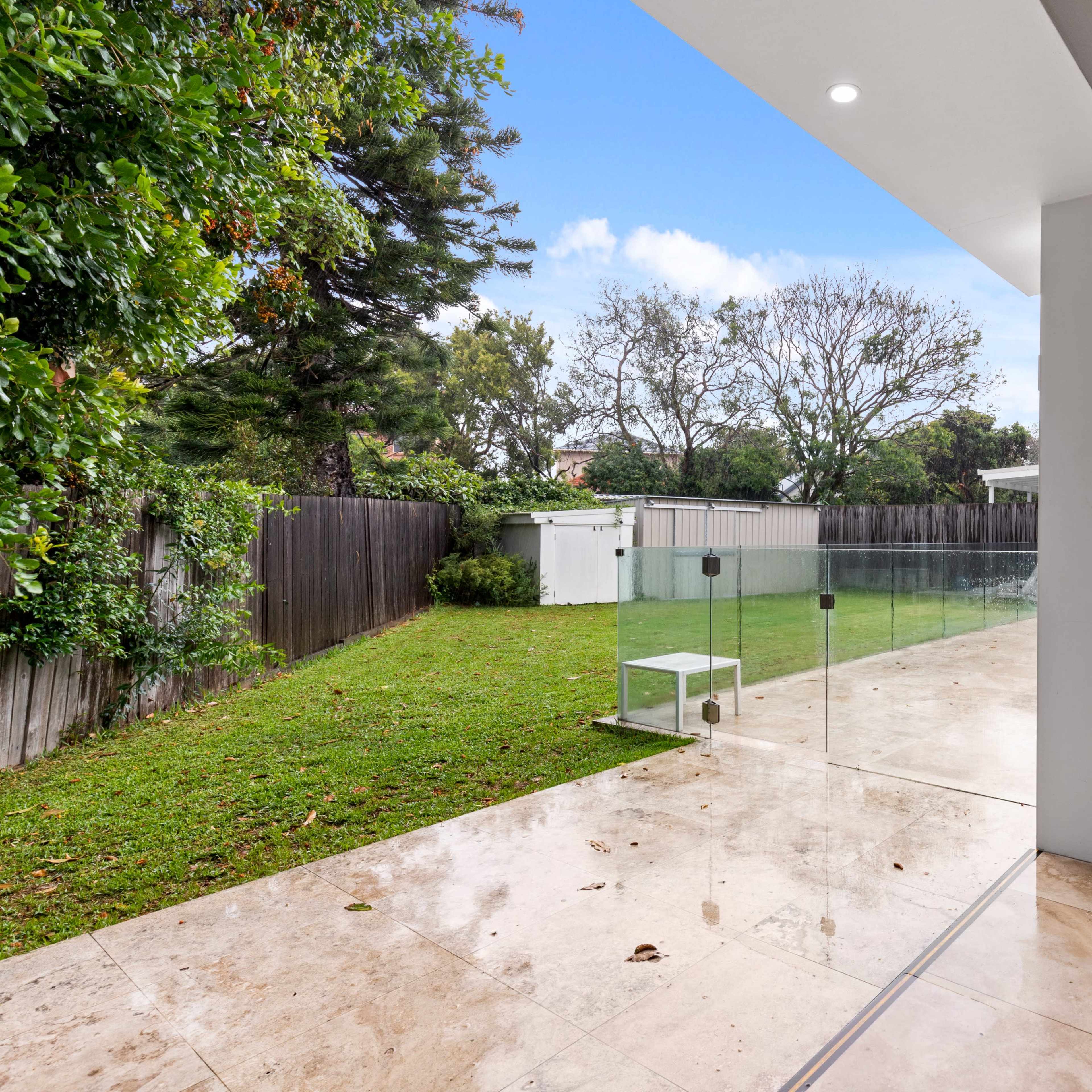 A modern backyard with a grassy area, a small shed, and a swimming pool, all bordered by a glass enclosure.
