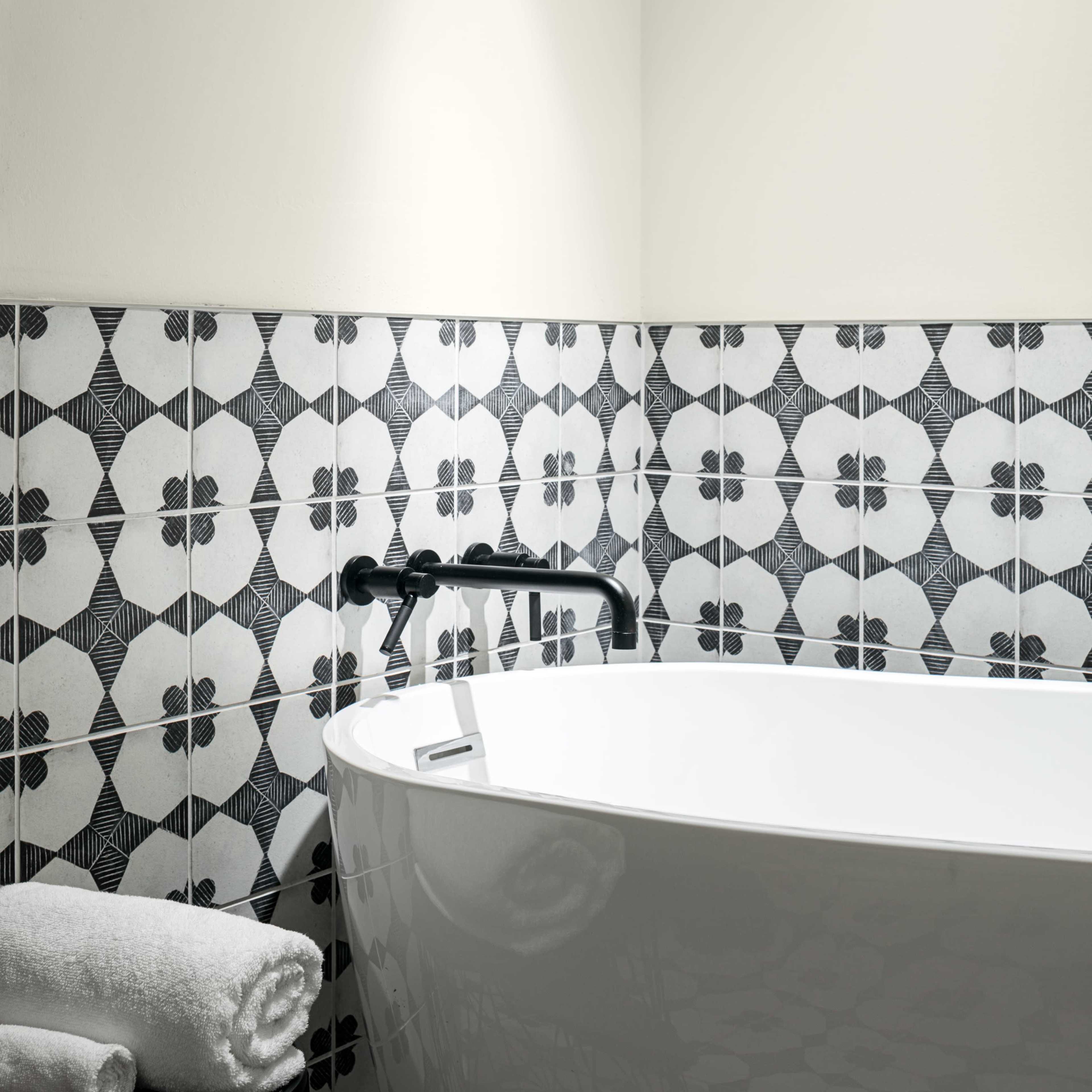 A modern bathroom featuring a white freestanding bathtub next to a black and white tiled wall and floor, with a small stool holding rolled towels.