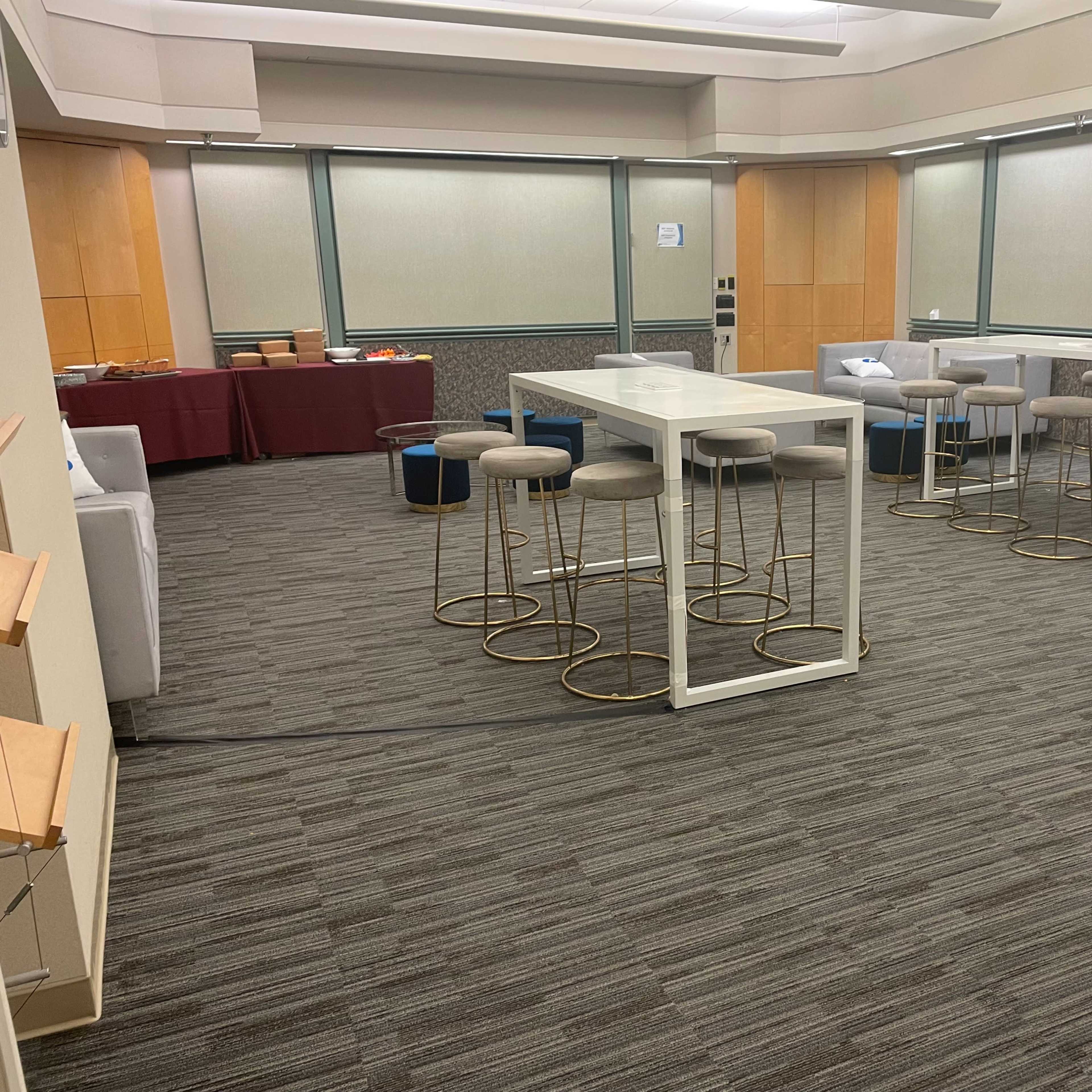 A spacious lounge area with a long table, several bar stools, and seating arrangements, complemented by light-colored walls and carpet flooring.