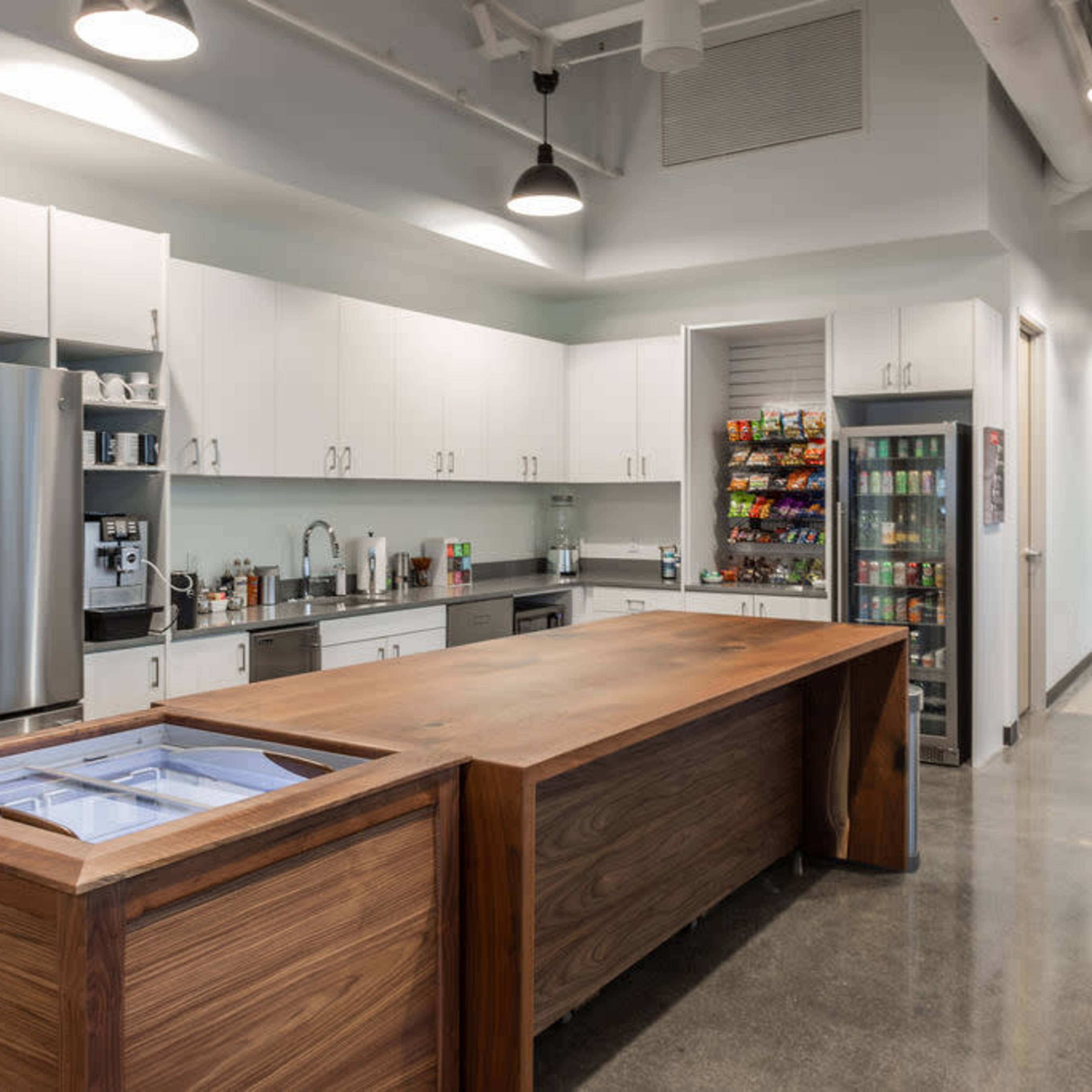 A modern kitchen area with a large wooden island, stainless steel appliances, and a vending machine in a bright workspace.