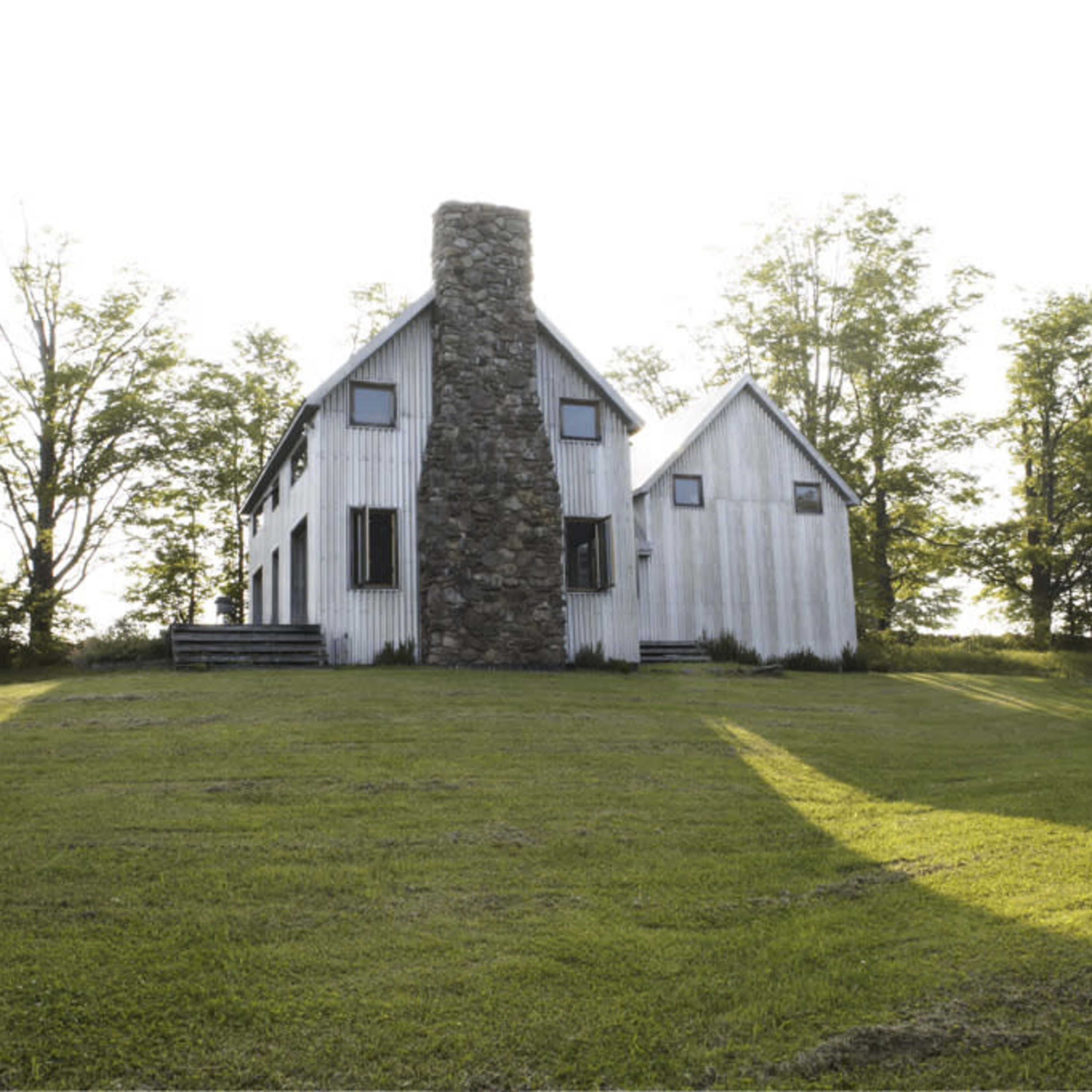 A two-story, white house with a stone chimney stands amidst a grassy area and trees.