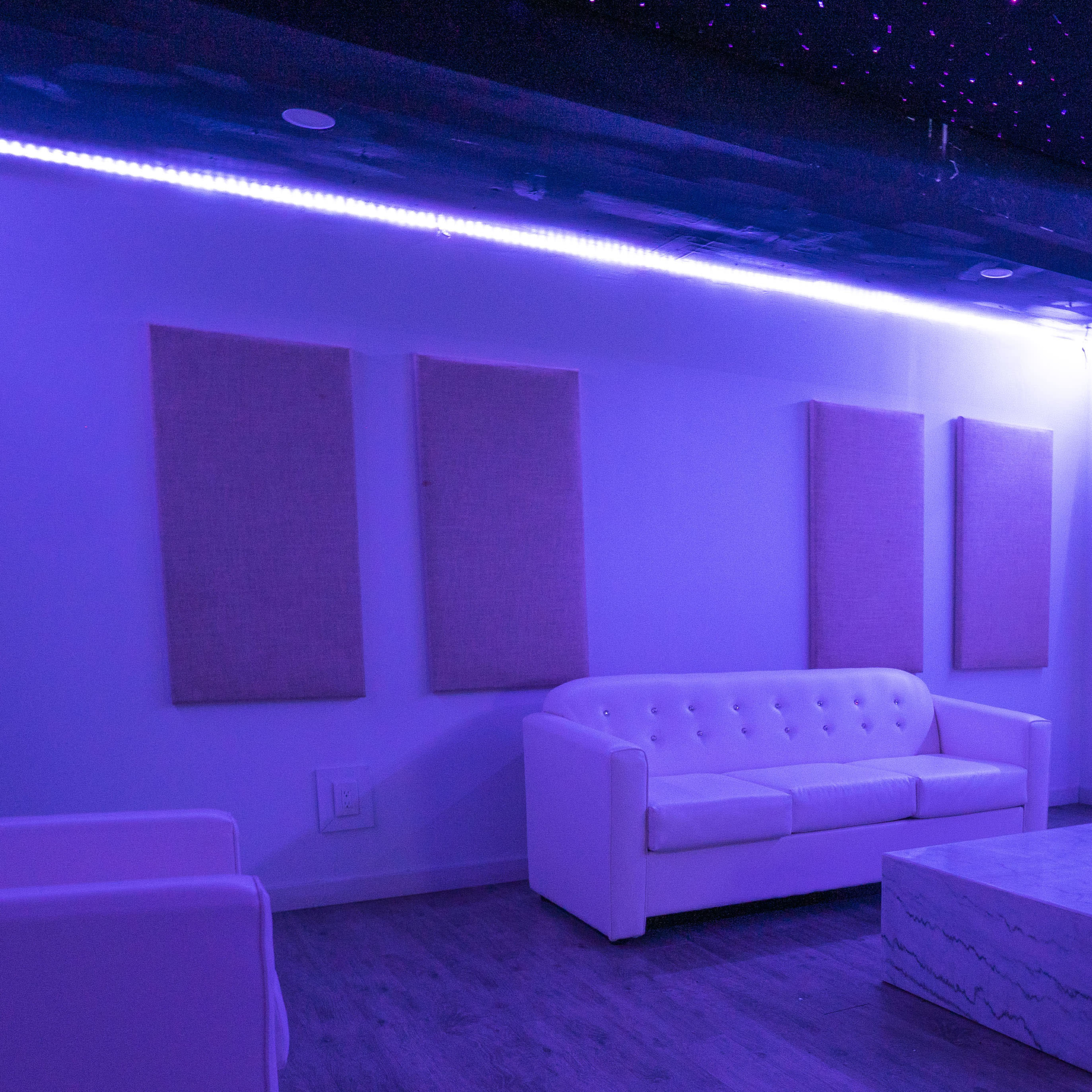 A lounge area with white furniture including a tufted couch and a marble coffee table, illuminated by purple LED lights.