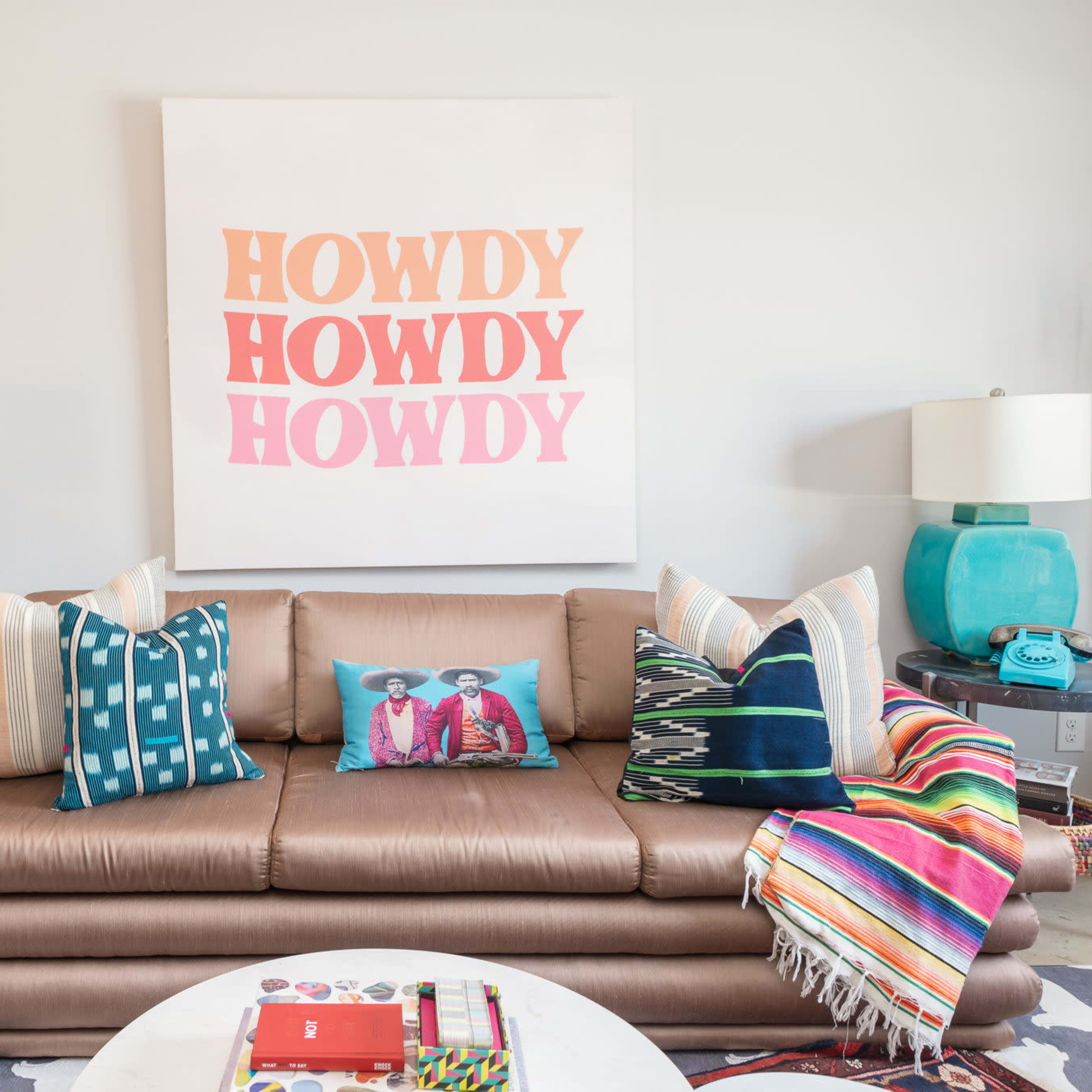 A modern living room features a brown sofa adorned with colorful pillows, a vibrant blanket draped over the side, and a large art piece with the word "HOWDY" displayed prominently above.