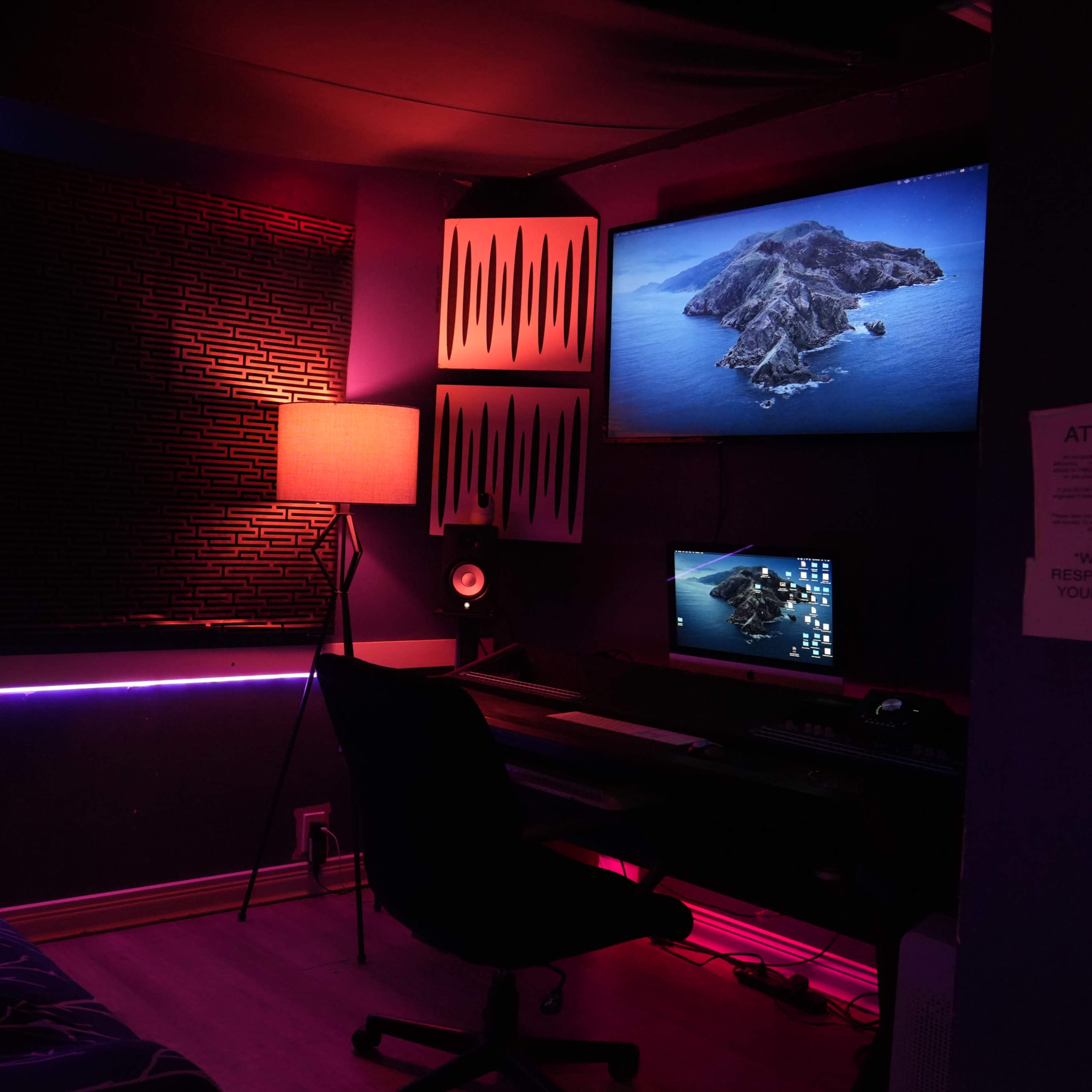 A small, dimly lit room with a desk setup, a computer monitor, and ambient lighting, including a lamp and LED strips.