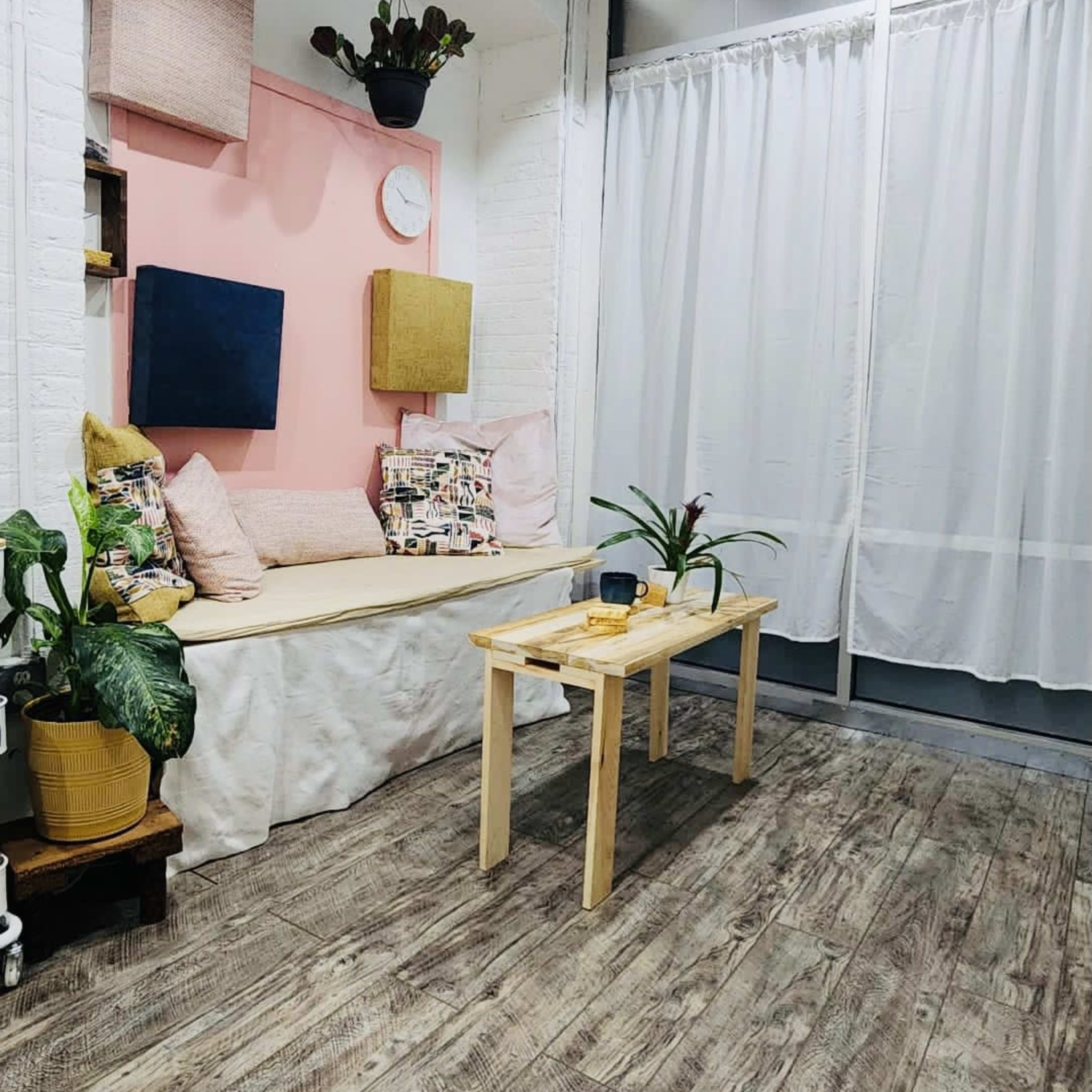 A cozy room with a seating nook against a pink wall, a small wooden table in the center, and plants in the corners.