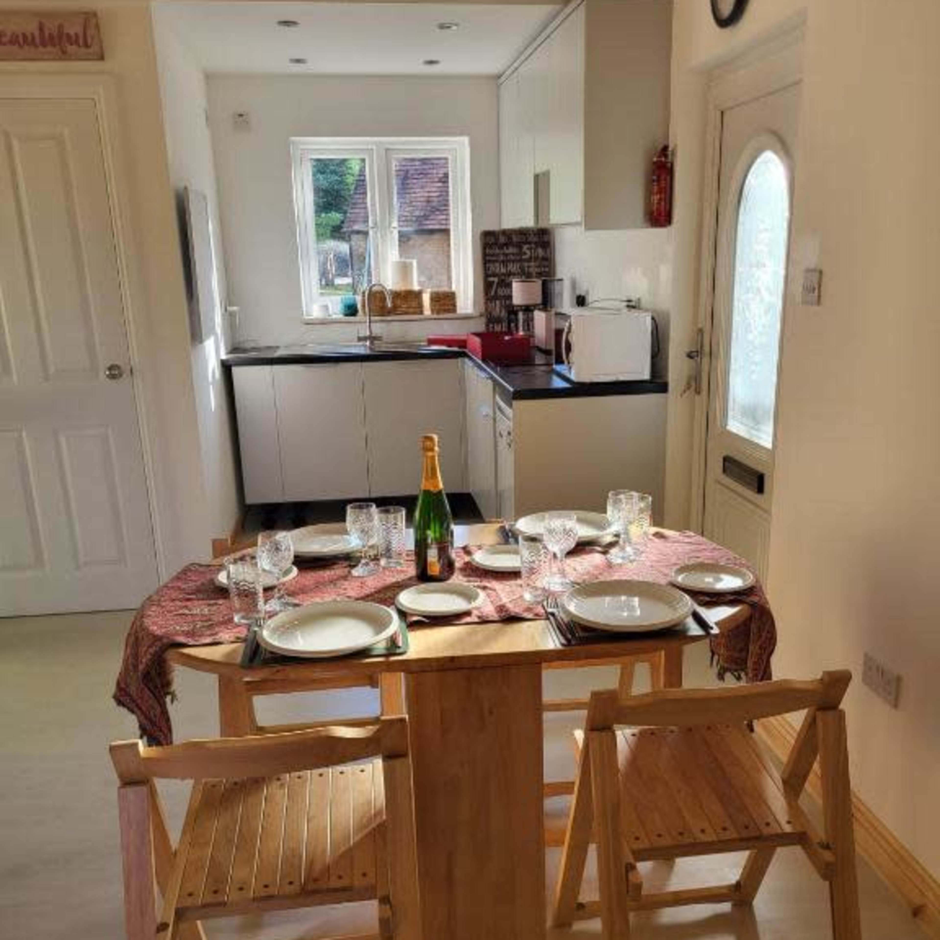 A round table set with plates and glasses is positioned in a kitchen area with cabinets and appliances in the background.