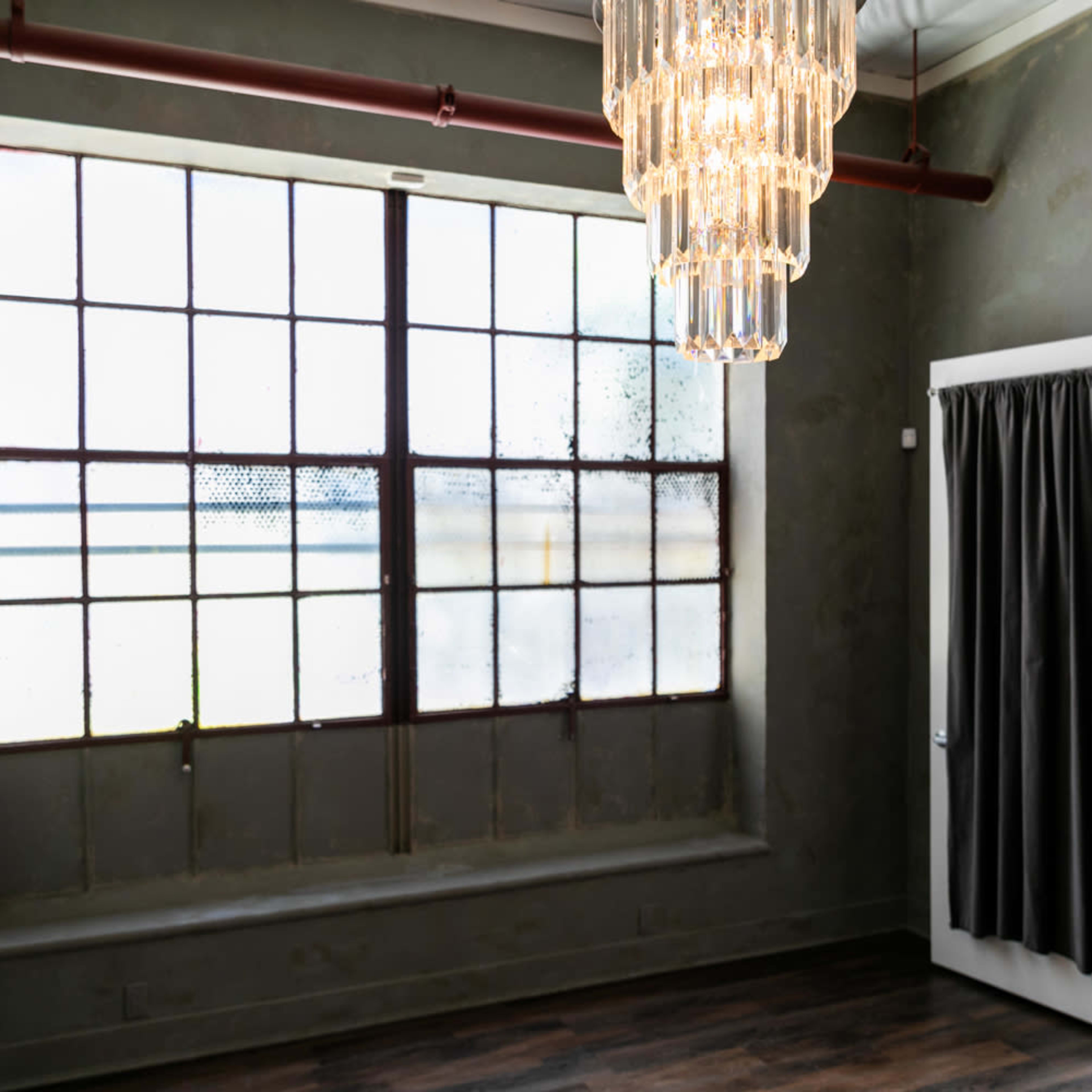 An industrial-style room with a large, divided window and a chandelier, complemented by an open door leading to another space.