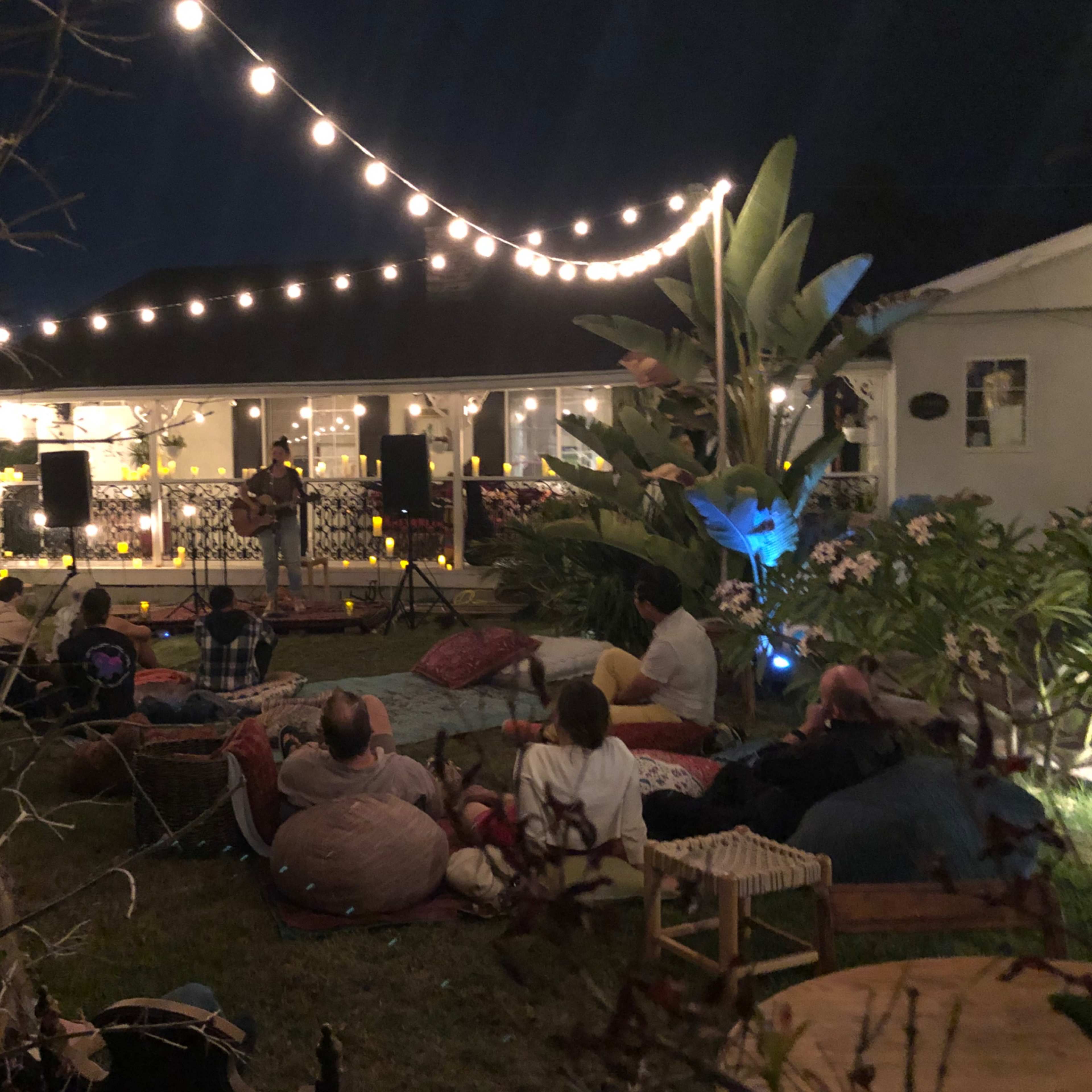 A group of people sits on blankets and cushions in a garden while a musician performs under string lights at night.