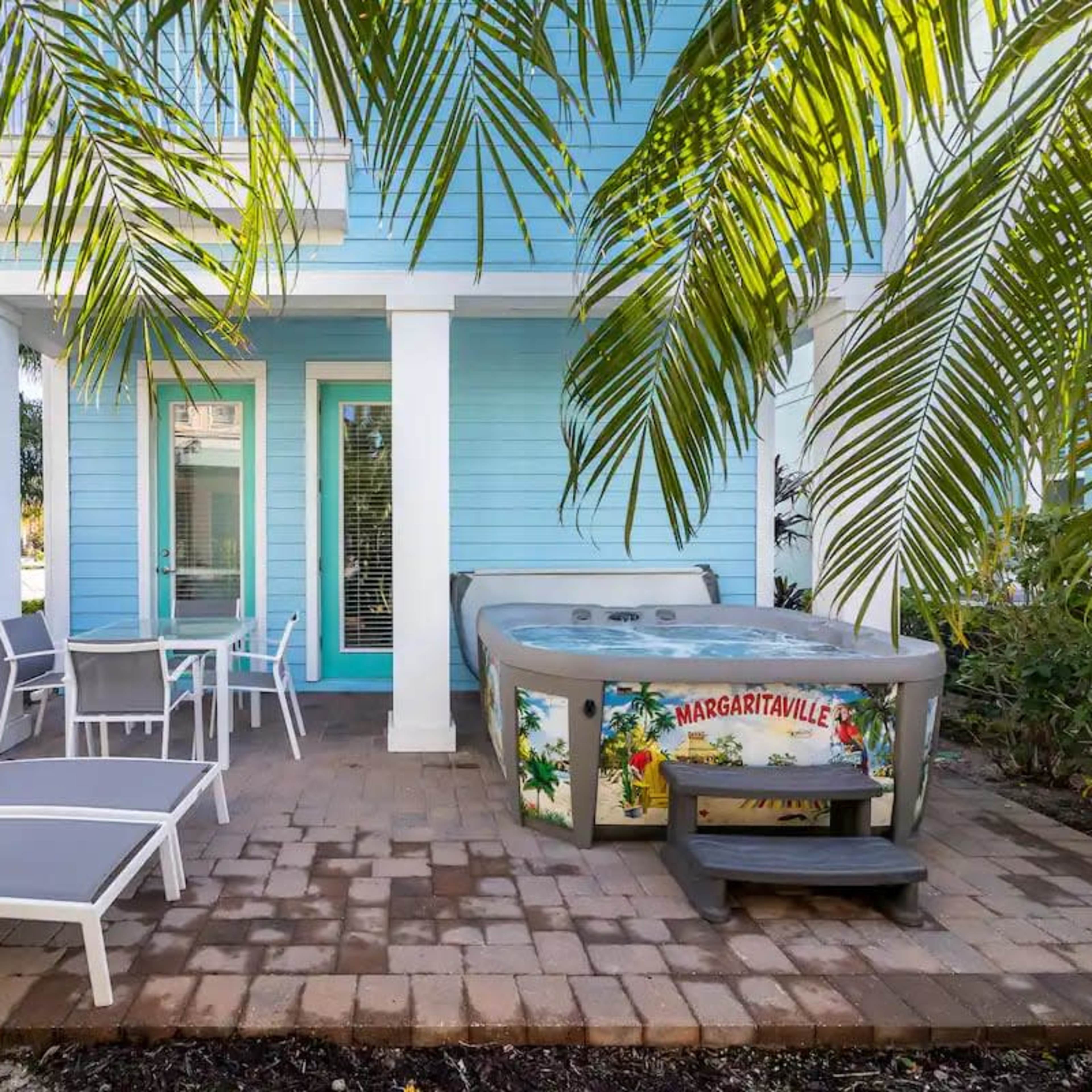 A hot tub is situated on a brick patio next to two lounge chairs and surrounded by tropical plants near a light blue house.