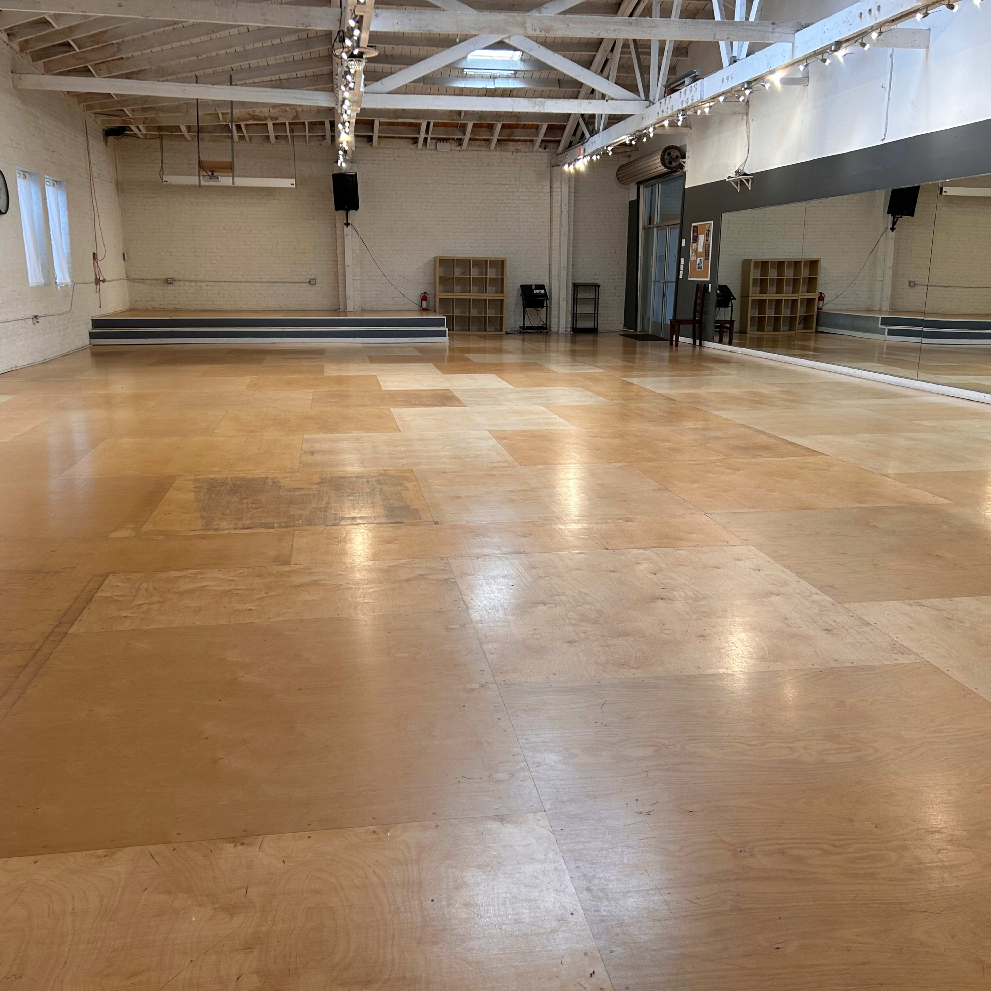 A spacious dance studio with wooden flooring, large mirrors along one wall, and natural light coming through several windows.