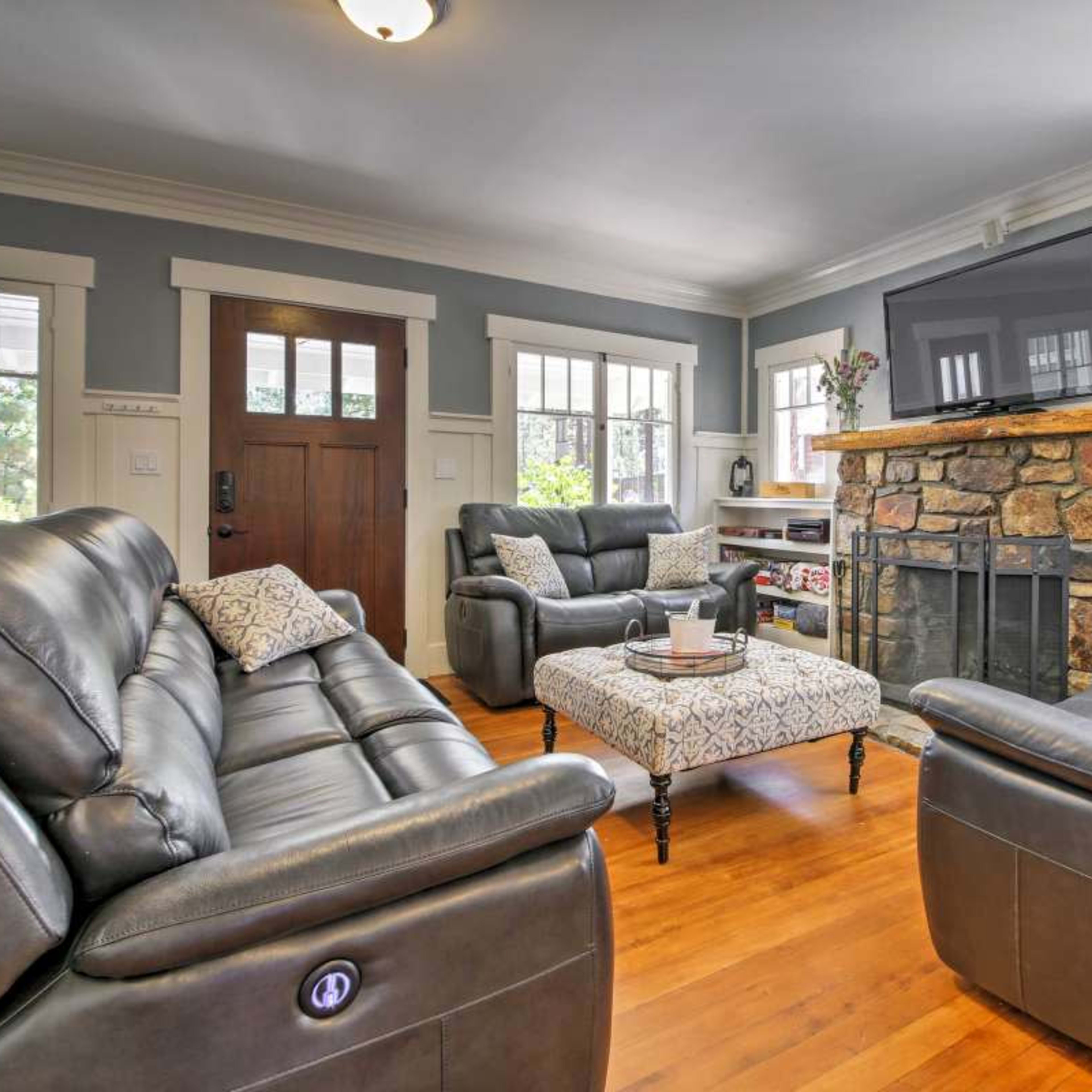 A living room featuring leather chairs and a couch, a stone fireplace, and a large television mounted on the wall, with wood flooring and natural light from several windows.