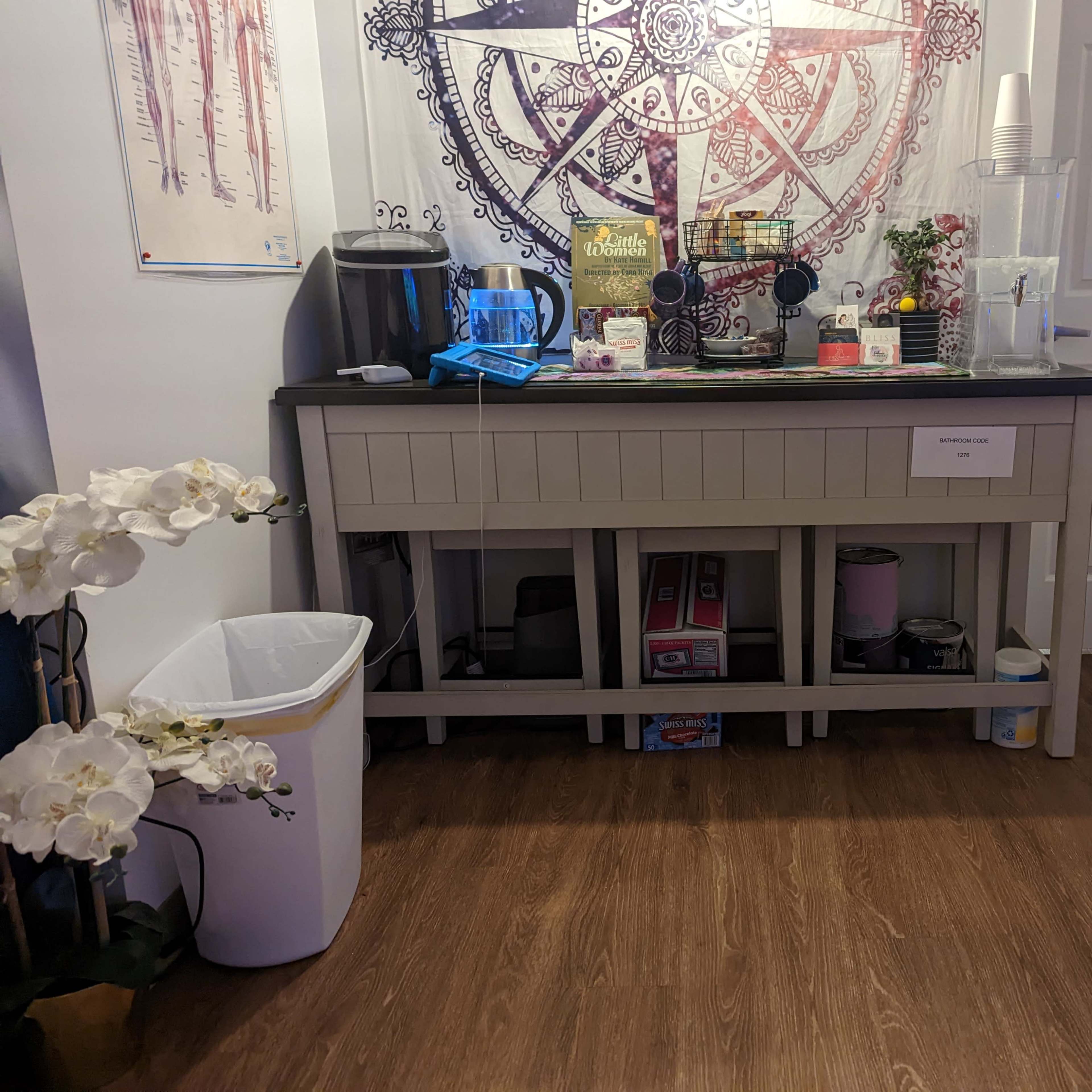 A beverage station with a counter, coffee machine, various drink supplies, and a trash bin, situated near a closed door and decorated with a large mandala.