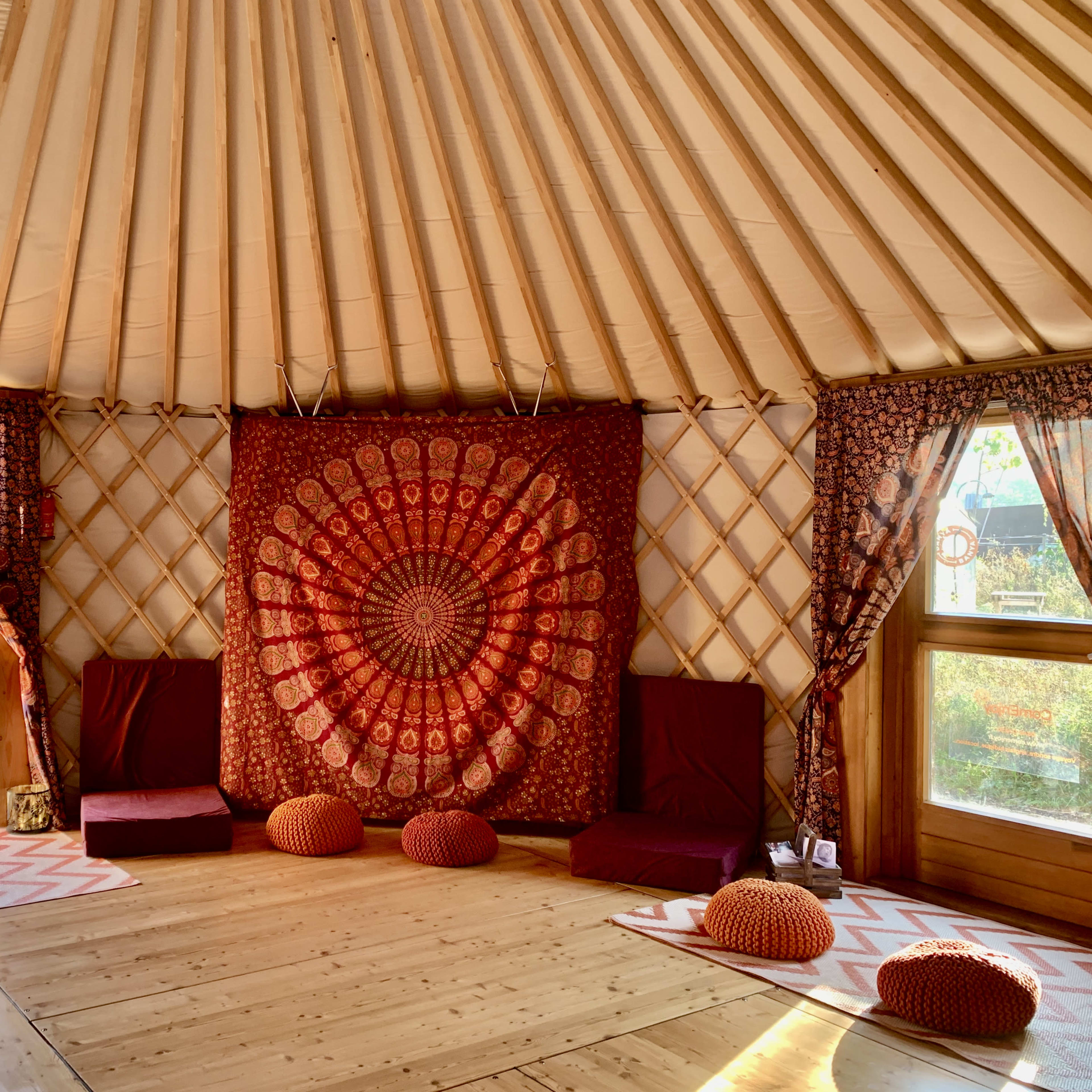 The interior of a yurt features a large circular tapestry, floor cushions, and wooden floors, with light coming through two windows adorned with patterned curtains.