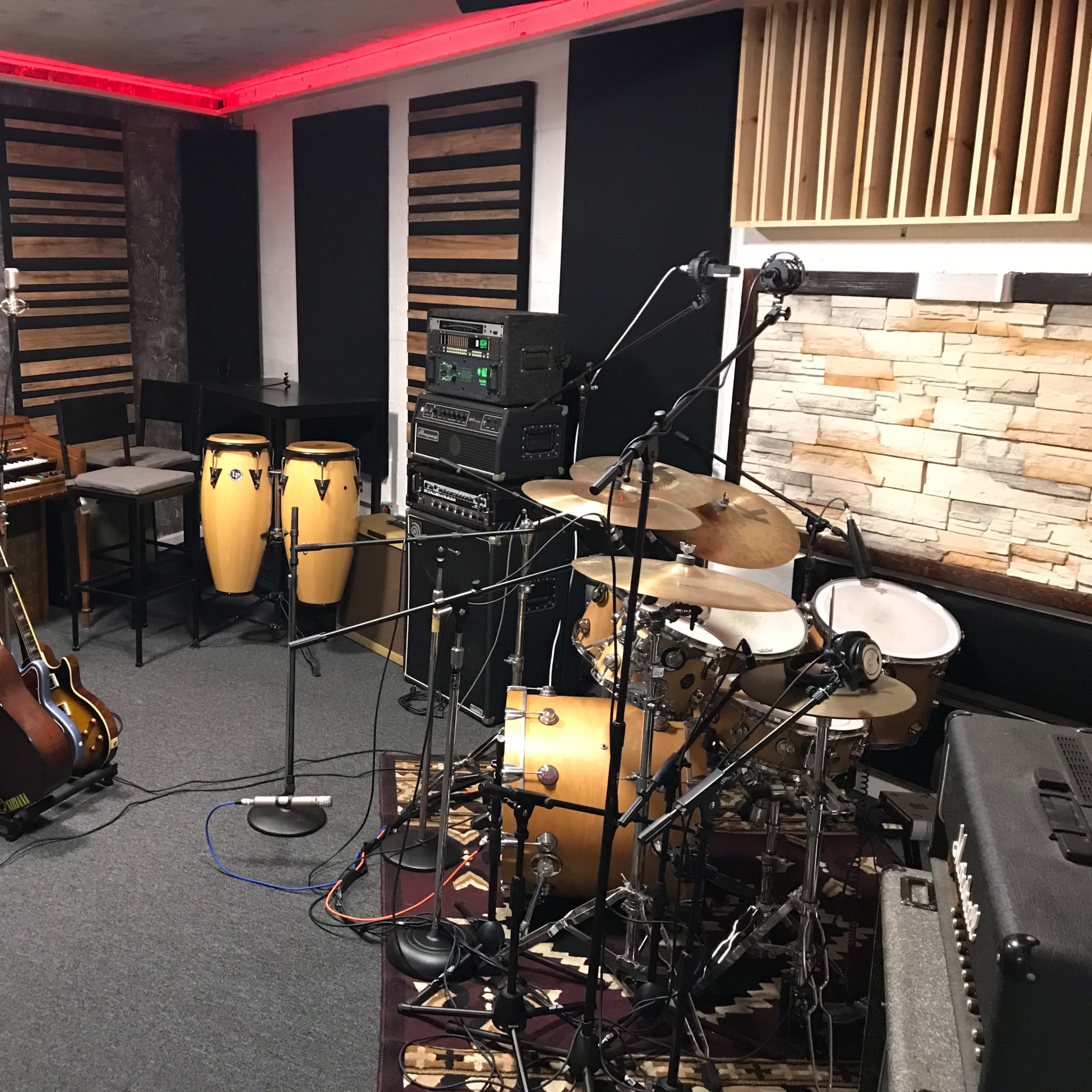 A music studio with various instruments, including a drum set, congas, an electric guitar on a stand, and a grand piano, surrounded by sound equipment and soundproofing materials.