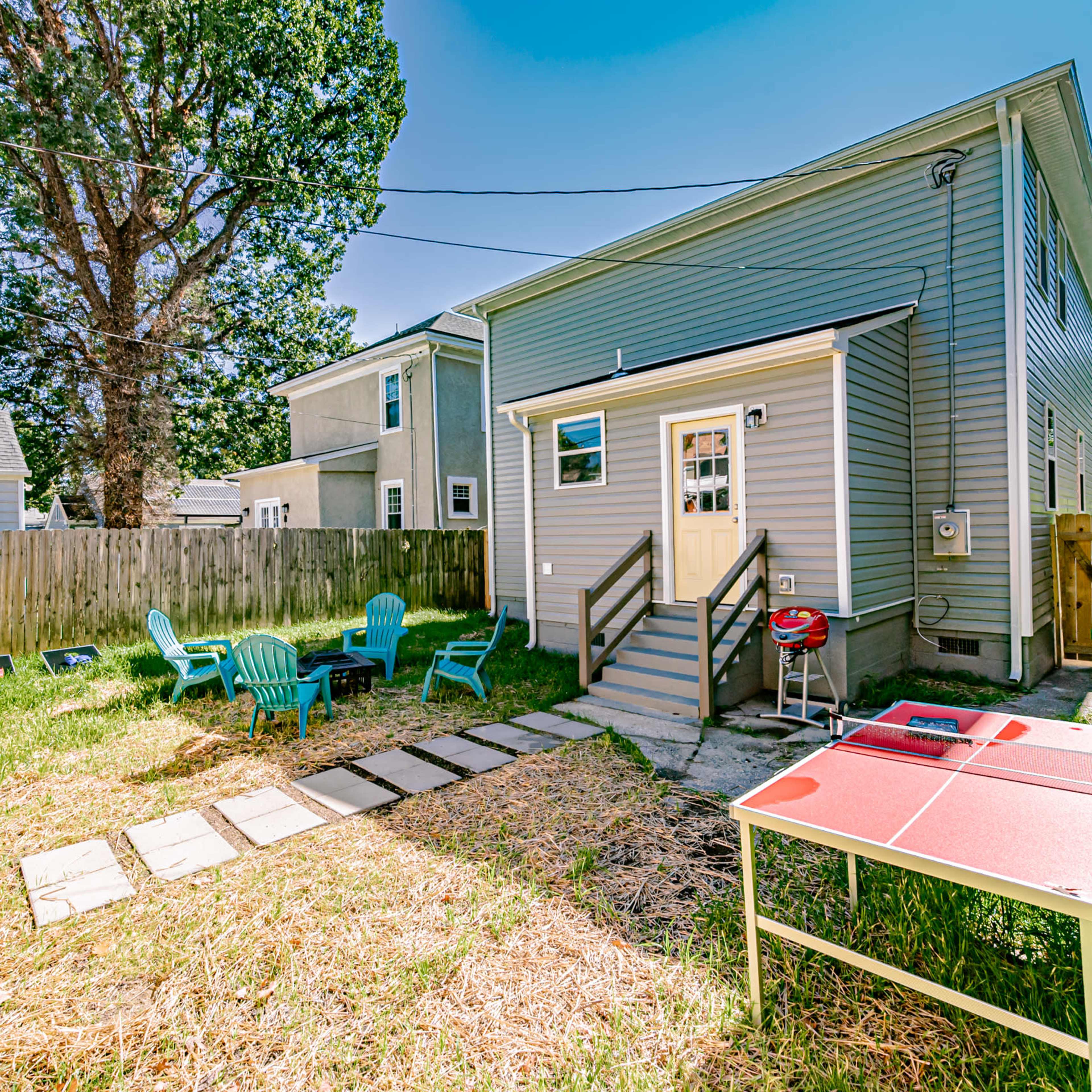 A backyard features a small patio area with green chairs, a barbecue grill, a ping pong table, and a pathway made of stone slabs.
