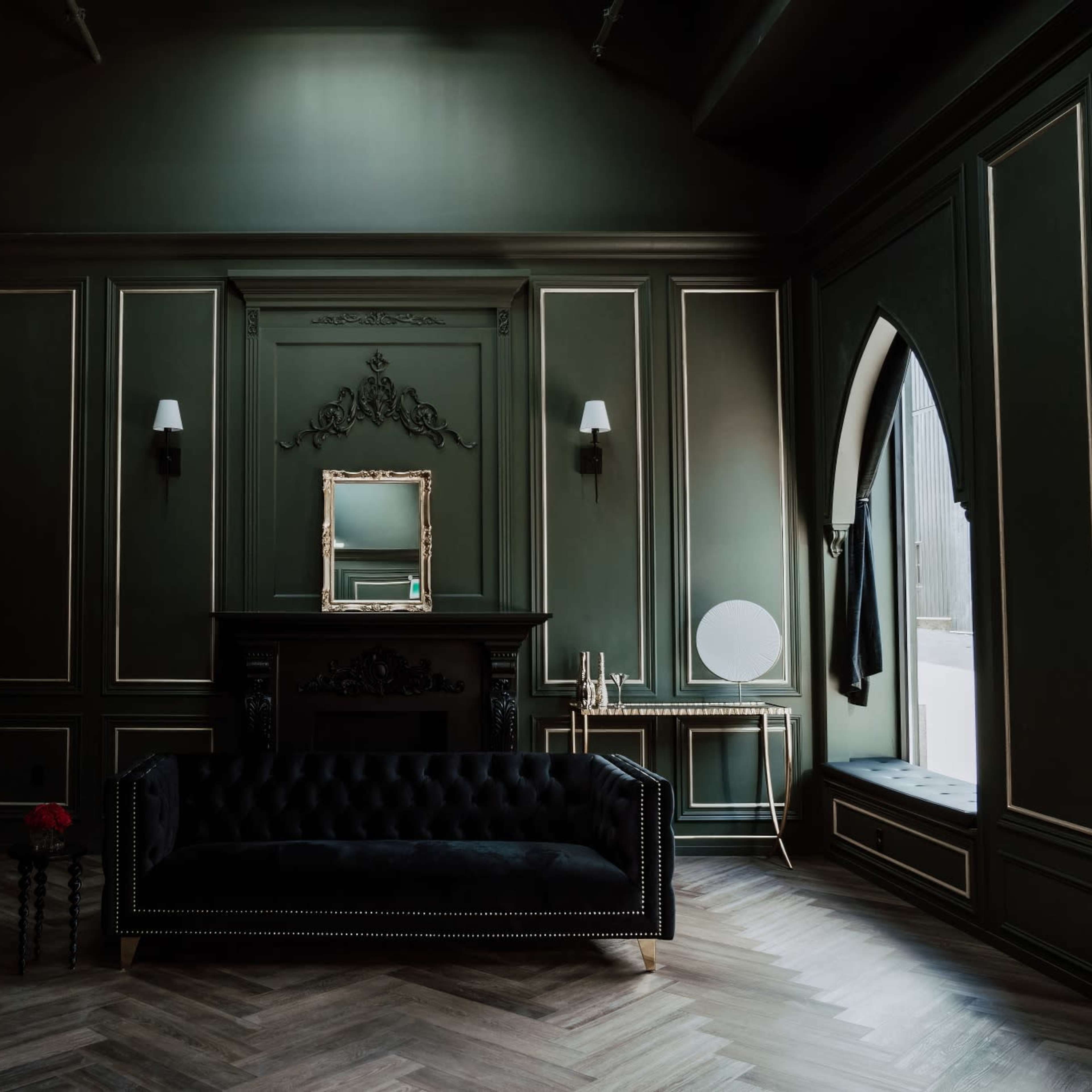 The room features dark green walls with ornate trim, a black velvet sofa, and a large window alongside bookshelves and decorative accents.