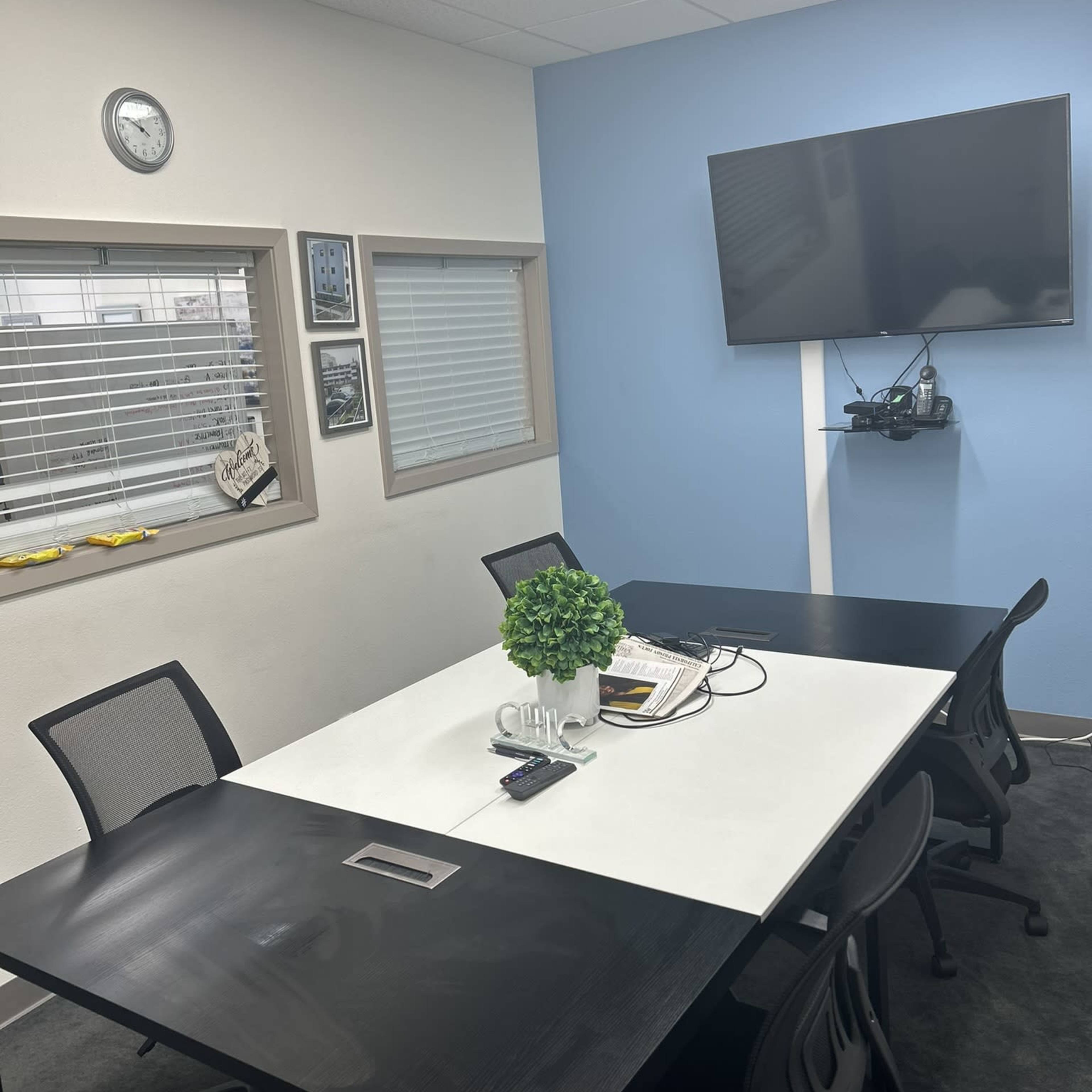 A conference room with a white table, black chairs, a wall-mounted television, and a plant centerpiece.