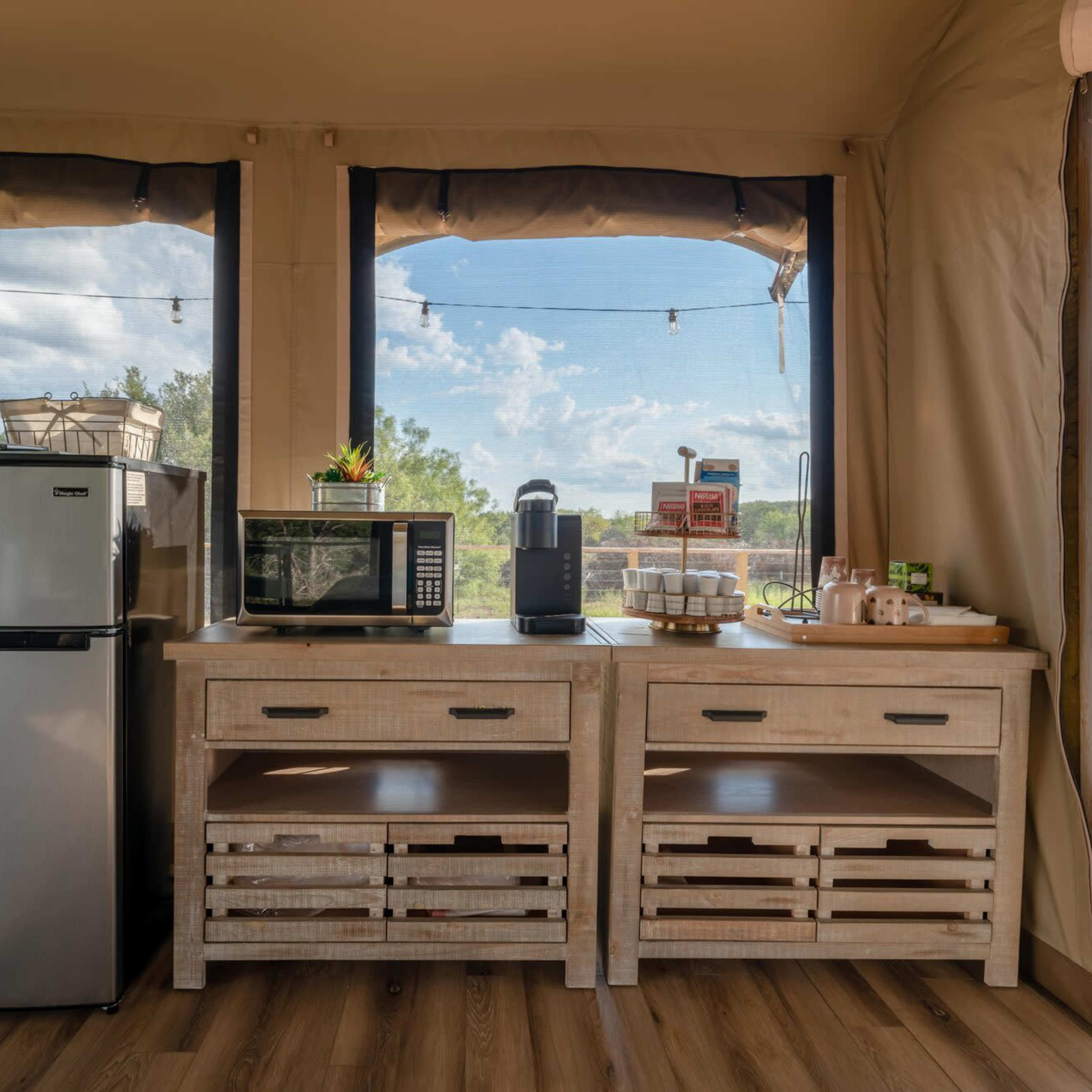 A cozy kitchen area with a mini refrigerator, microwave, and a coffee station, situated in a tent-like structure with large windows overlooking greenery.