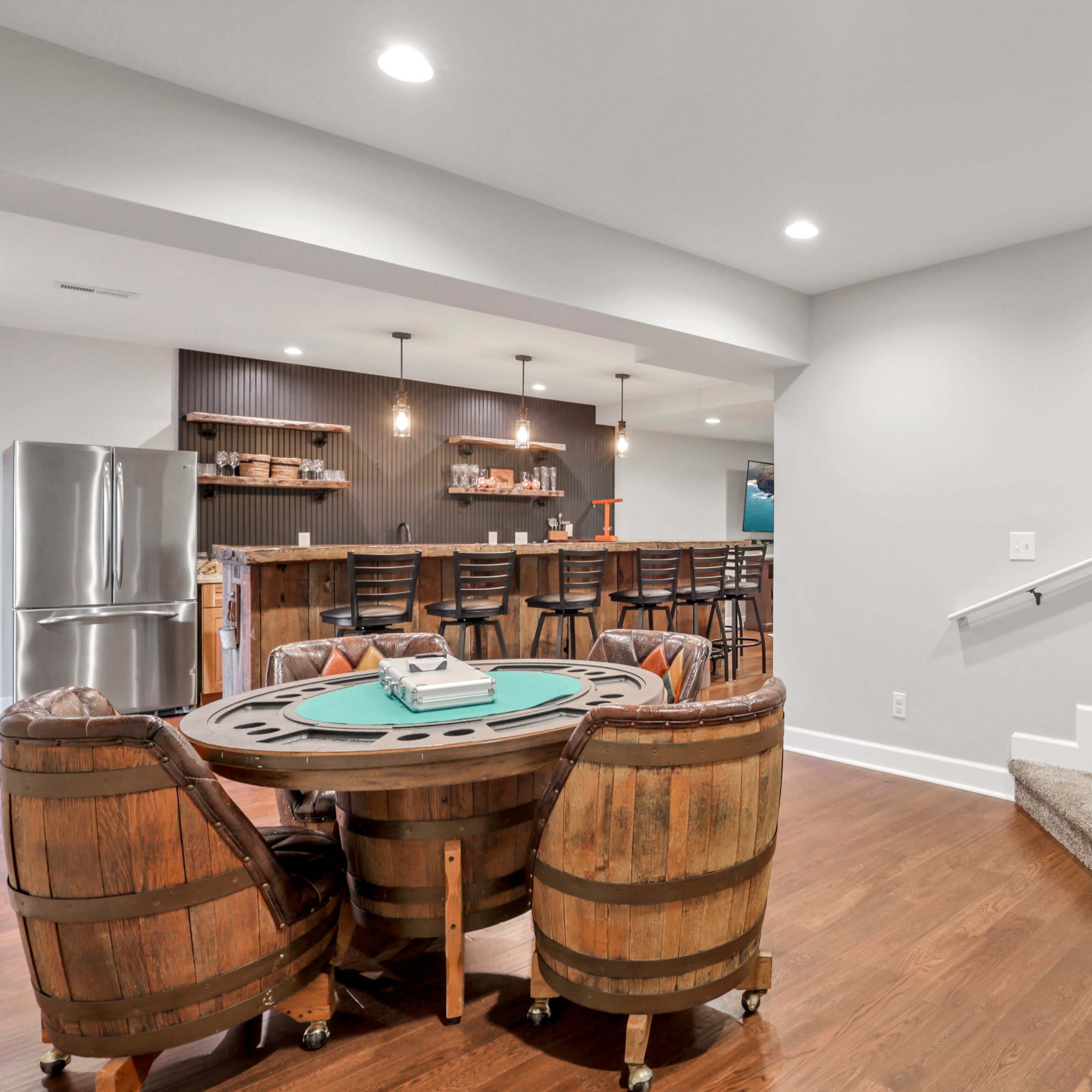 A basement interior featuring a poker table surrounded by wooden barrel chairs, a kitchenette bar in the background, and a staircase leading to an upper level.