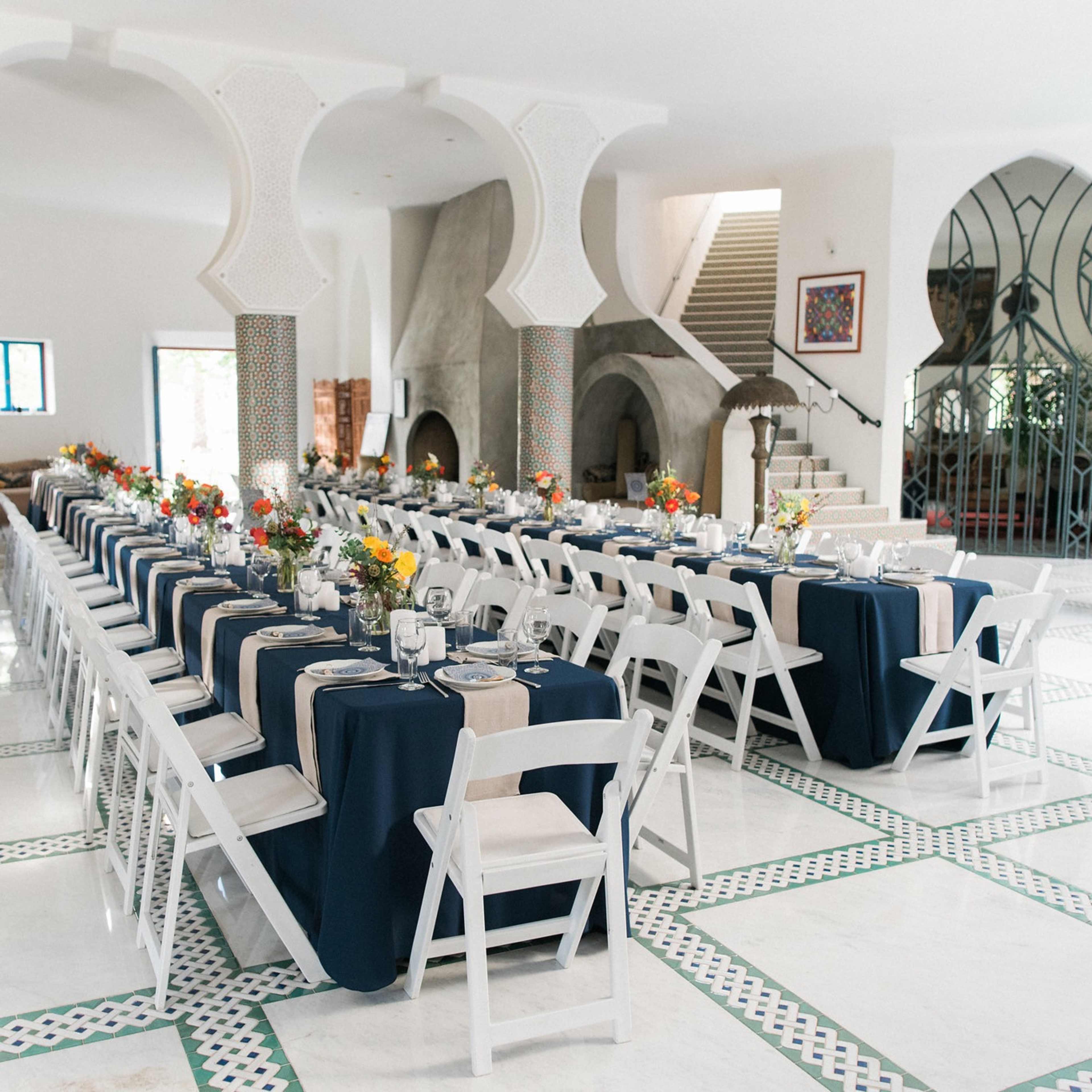 A spacious dining area features long tables set with navy blue tablecloths, white chairs, and floral centerpieces in a well-lit, modern room with arched columns.